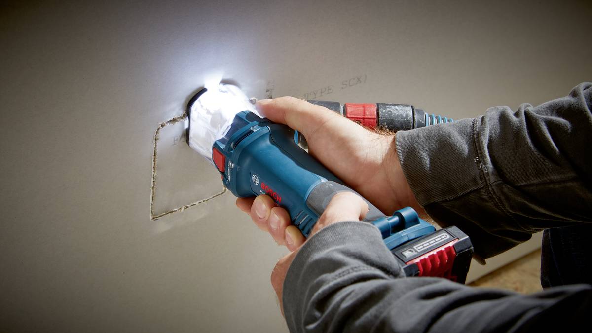 Une personne utilise un appareil électrique bleu pour découper un trou rectangulaire dans une cloison en plaques de plâtre.
