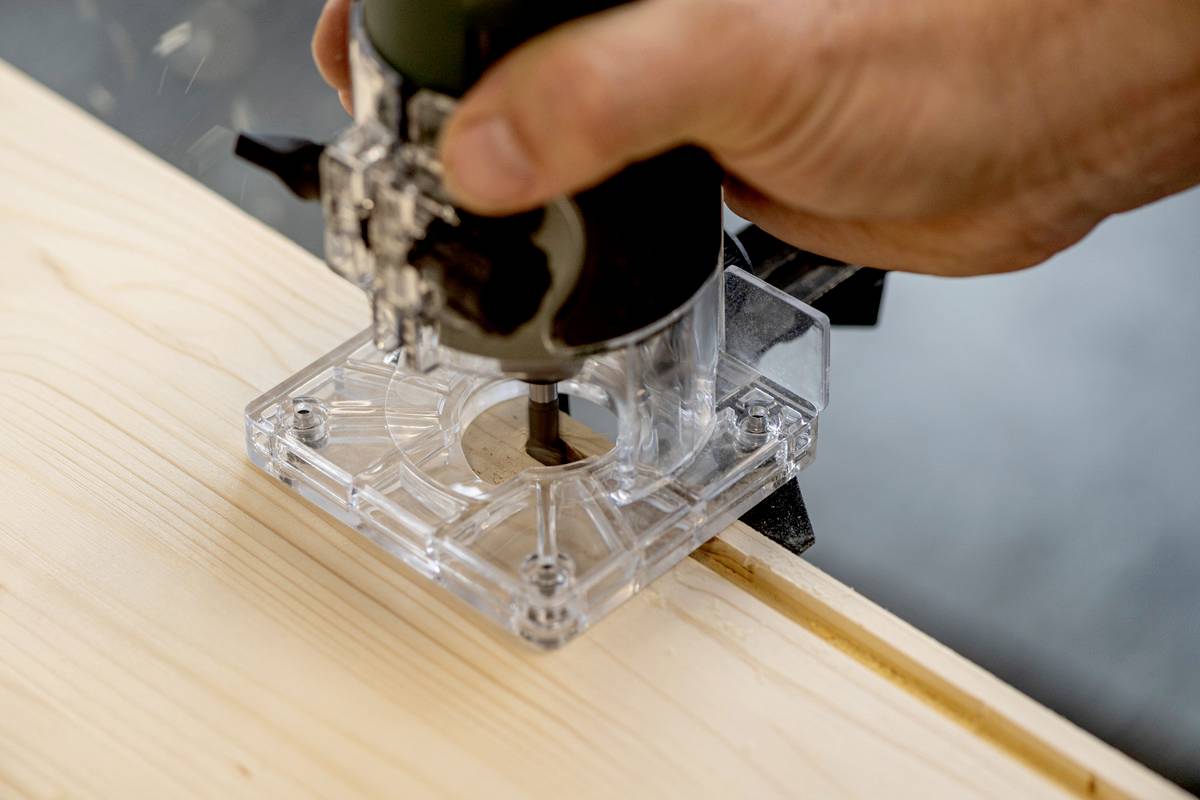 Une personne utilise une défonceuse sur une planche de bois. La fraiseuse est transparente et contribue à l'usinage du bois.