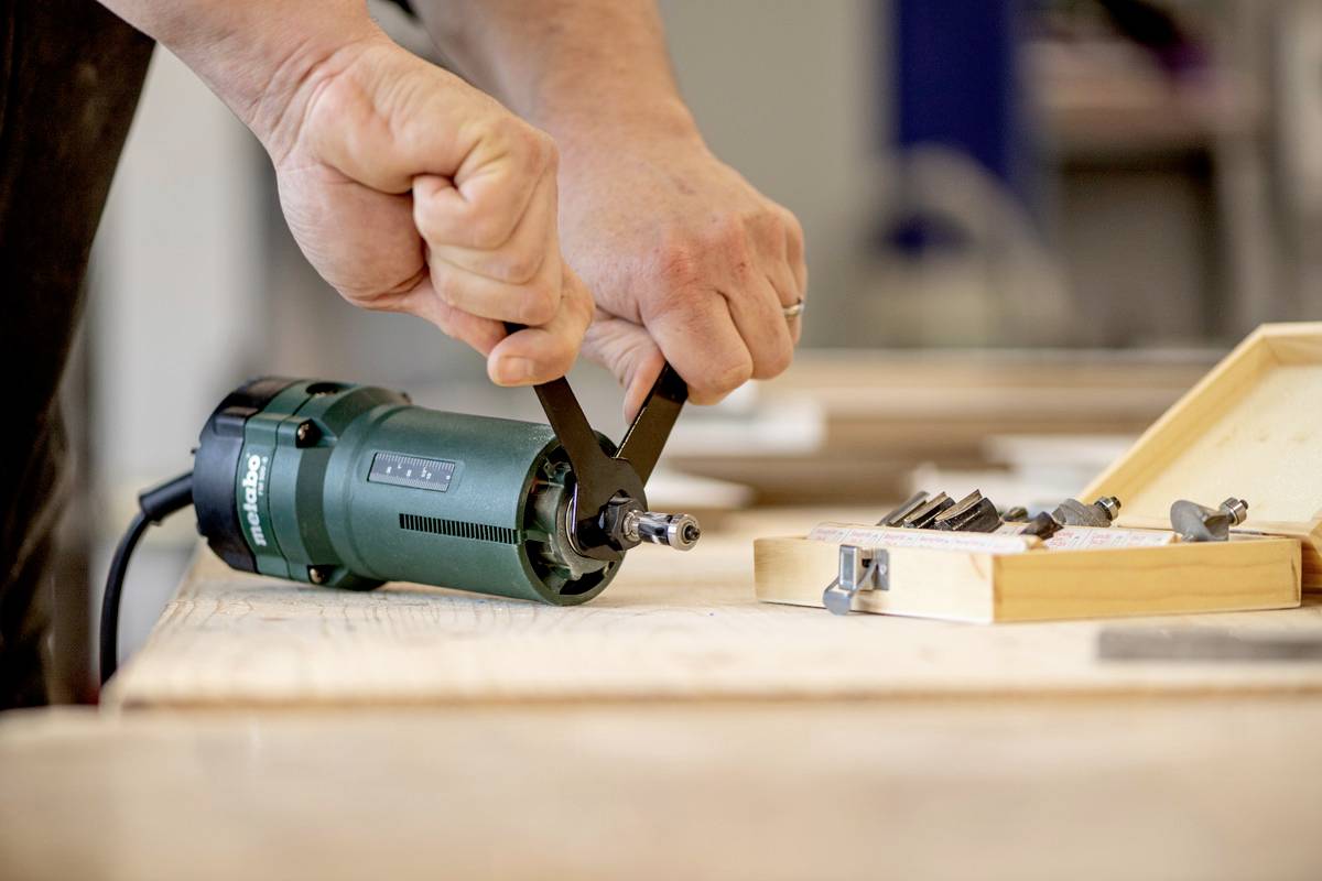 Gros plan sur des mains en train de monter un outil. Une perceuse verte est posée sur une table en bois, à côté d'un coffret en bois ouvert contenant des embouts.