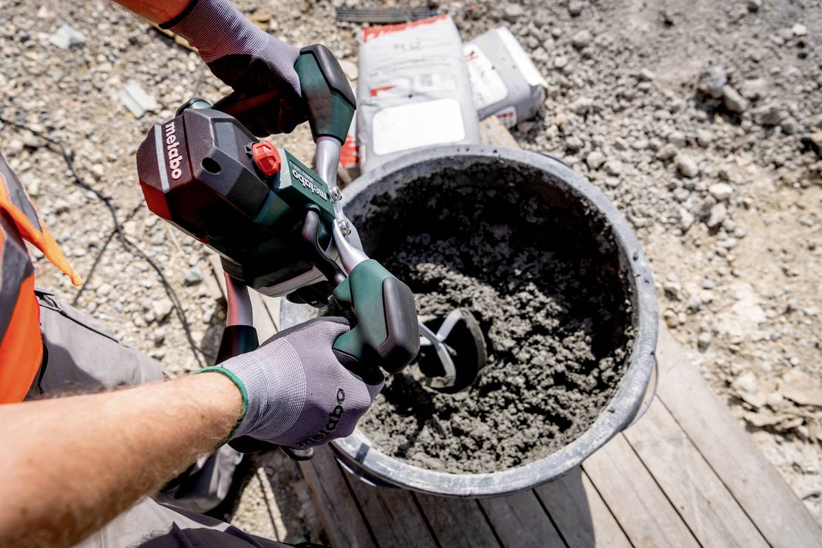Une personne mélange du béton dans un seau avec un mélangeur électrique à main. Le sol est rocailleux, et des matériaux de construction sont disposés à proximité.