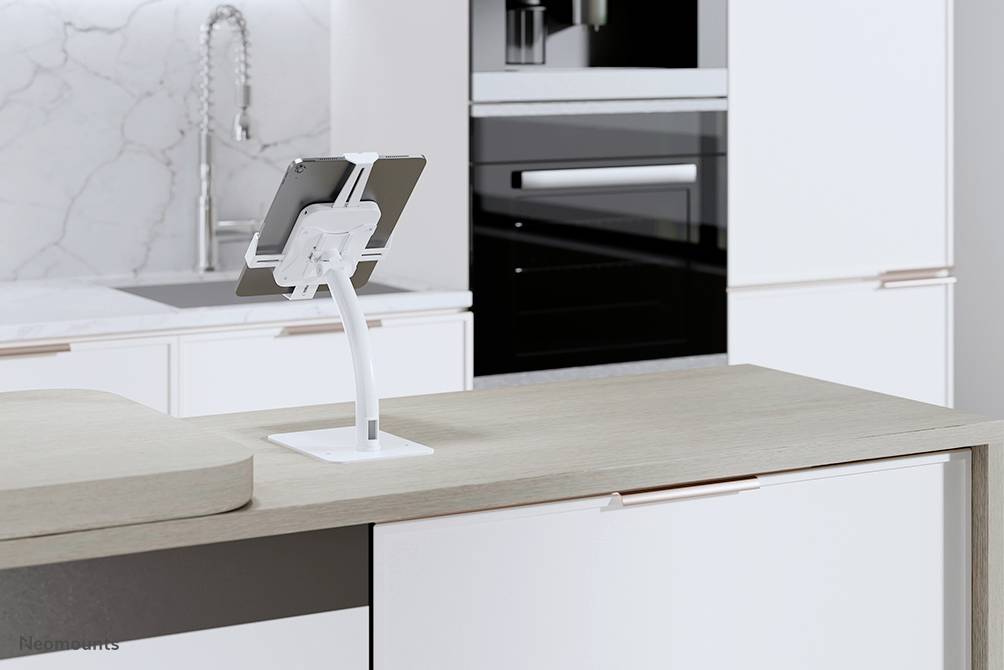 Une tablette sur un support blanc posée sur un plan de travail de cuisine moderne. L'arrière-plan montre des armoires blanches et un robinet.