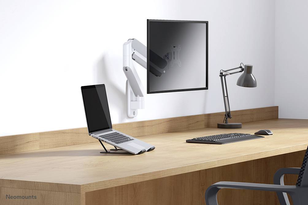 Un bureau avec un ordinateur portable sur un support à côté d'un écran monté au mur. Une lampe et un clavier sont également présents.