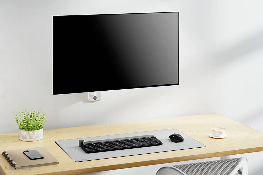 Un bureau bien rangé avec un moniteur mural, un clavier et une souris sur un tapis de souris gris, un carnet de notes, un stylo et une plante en pot.