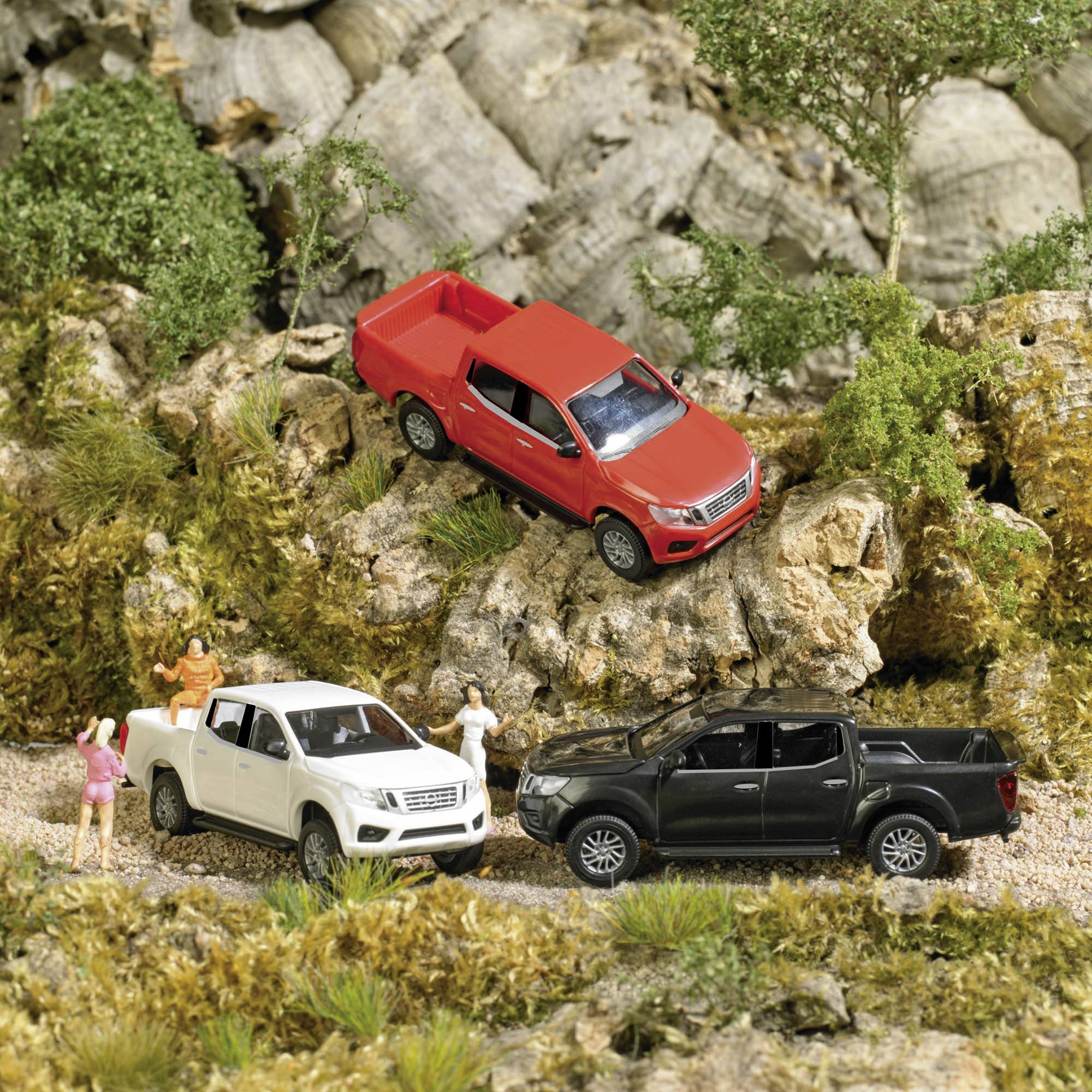 Trois véhicules miniatures (rouge, blanc, noir) sont disposés dans un paysage miniature avec des rochers et des arbres. Des figurines humaines observent la scène.