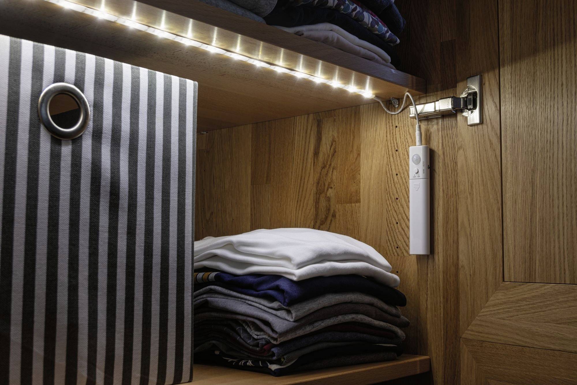 Un placard en bois avec des vêtements pliés sur une étagère et un conteneur rayé. Une bande LED éclaire l'intérieur.