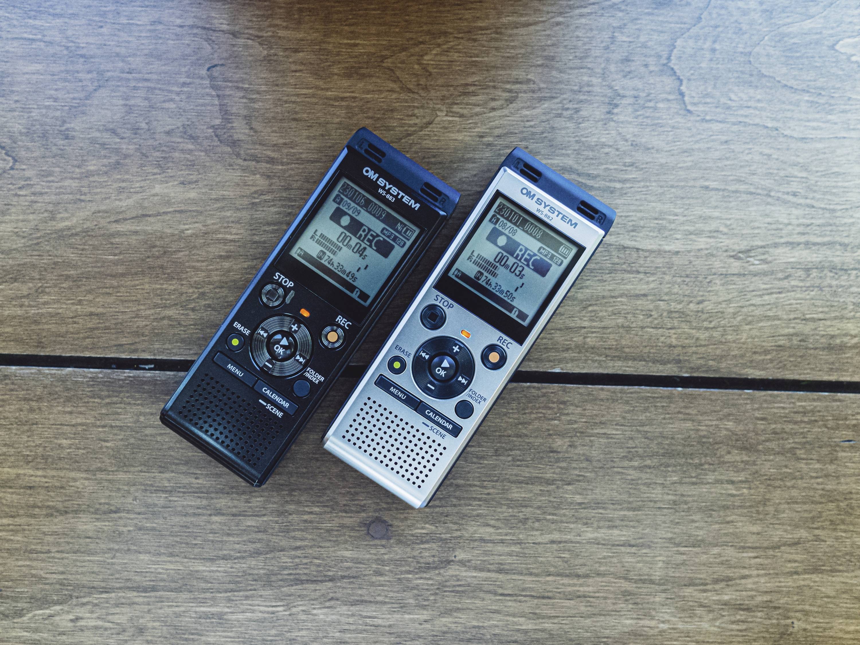 Deux dictaphones sont posés côte à côte sur une table en bois. L'un est noir, l'autre argenté. Tous deux présentent un écran LCD affichant le temps d'enregistrement.