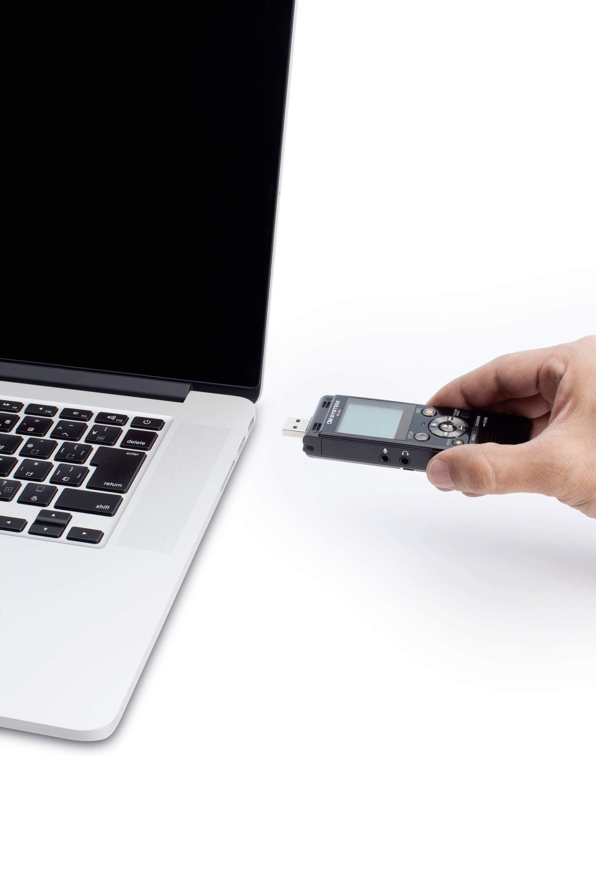 Une main tient un dictaphone qui est connecté à un ordinateur portable. L'ordinateur portable affiche un écran noir et possède un boîtier argenté.