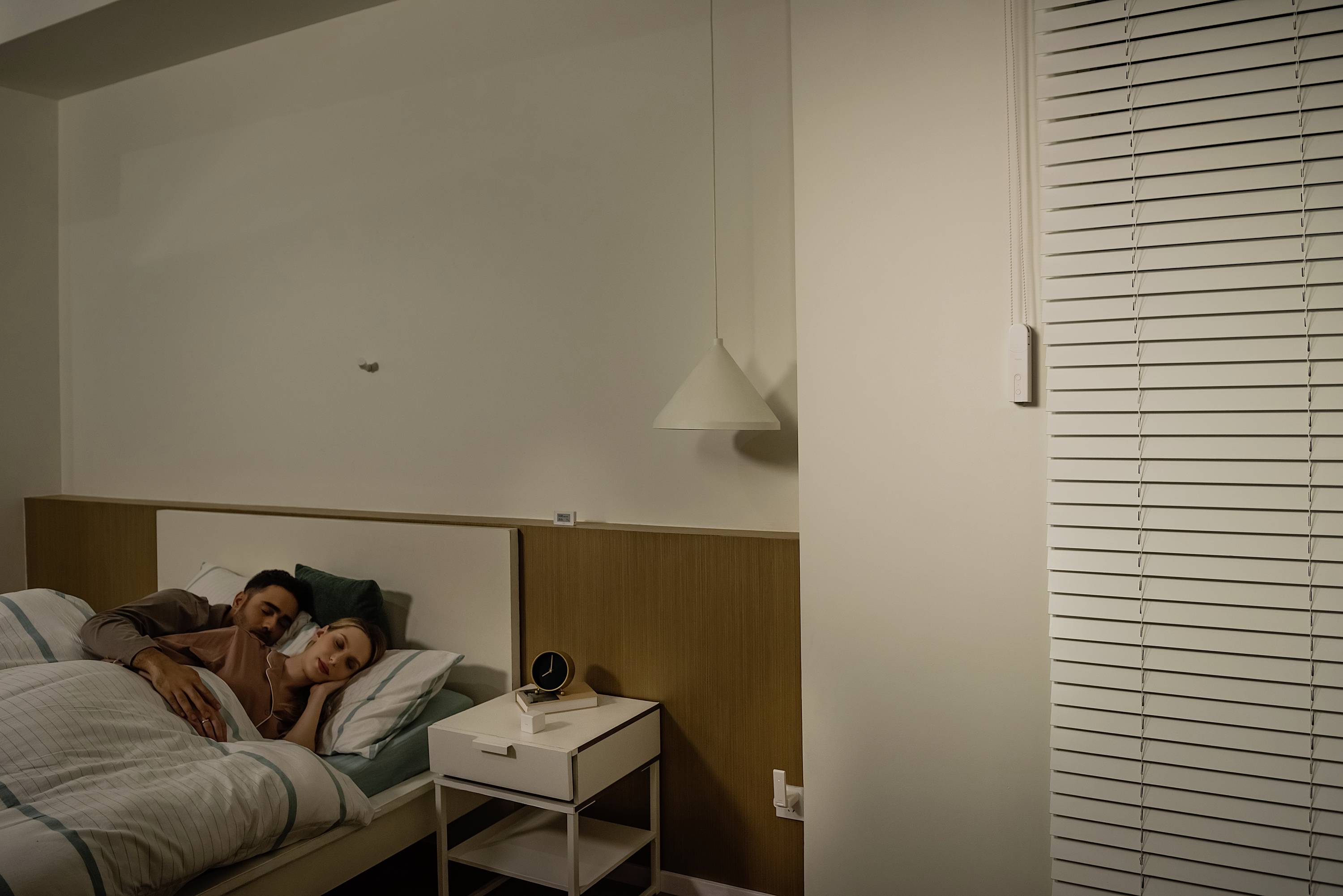 Un couple est confortablement allongé dans un lit, dans une chambre à l'éclairage tamisé. Une table de chevet avec une lampe se trouve à côté du lit.