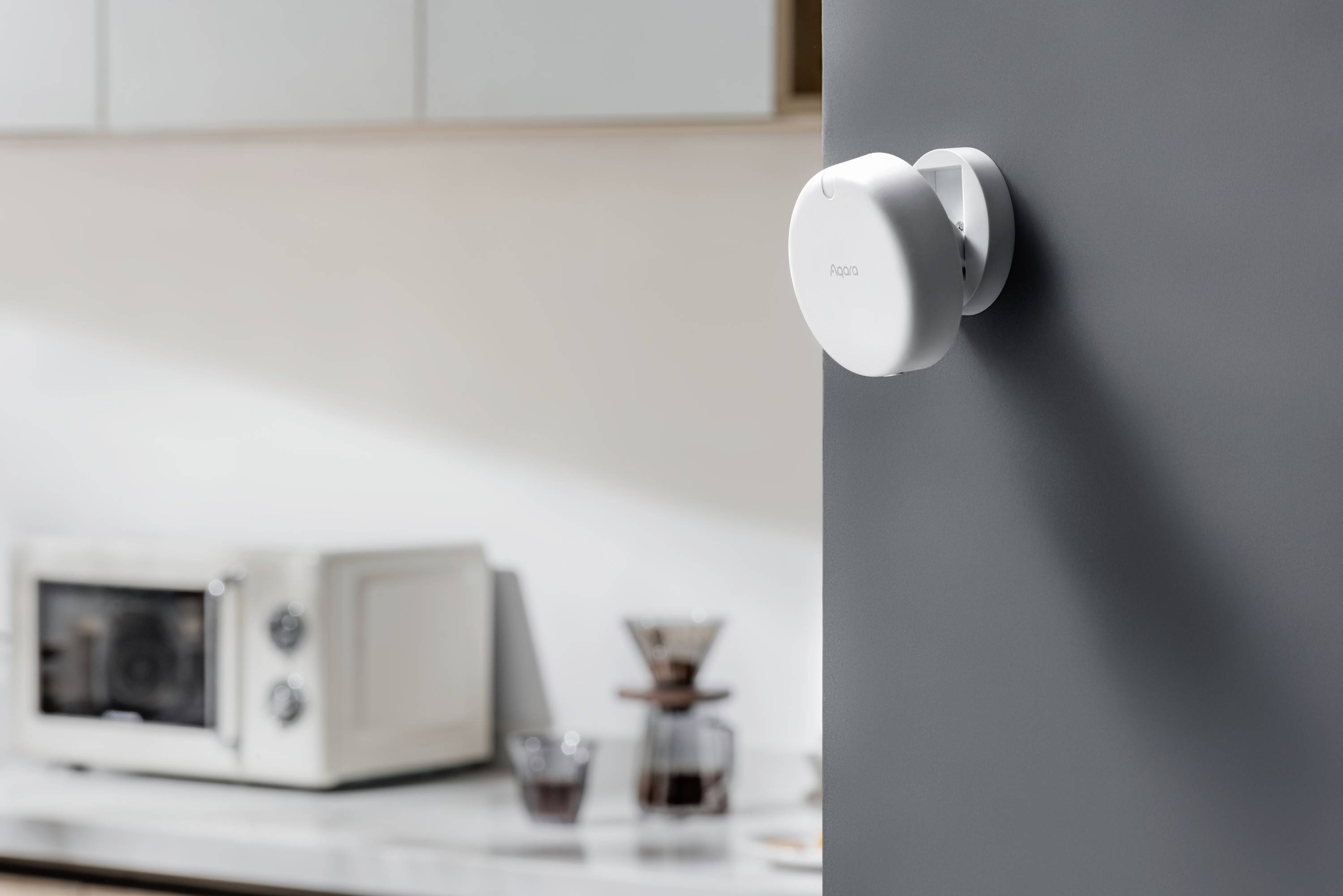 Un thermostat de forme ronde et moderne fixé sur un mur gris dans une cuisine. Une micro-ondes et une machine à café sont visibles en arrière-plan.