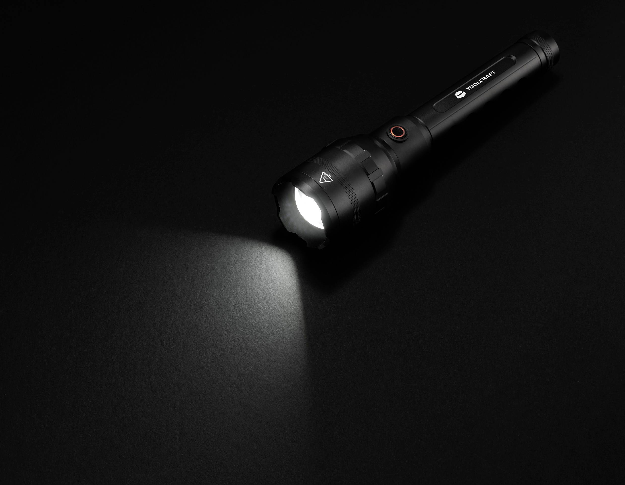 Une lampe de poche noire est posée sur un fond sombre. Elle est allumée et projette un faisceau lumineux vers la gauche.