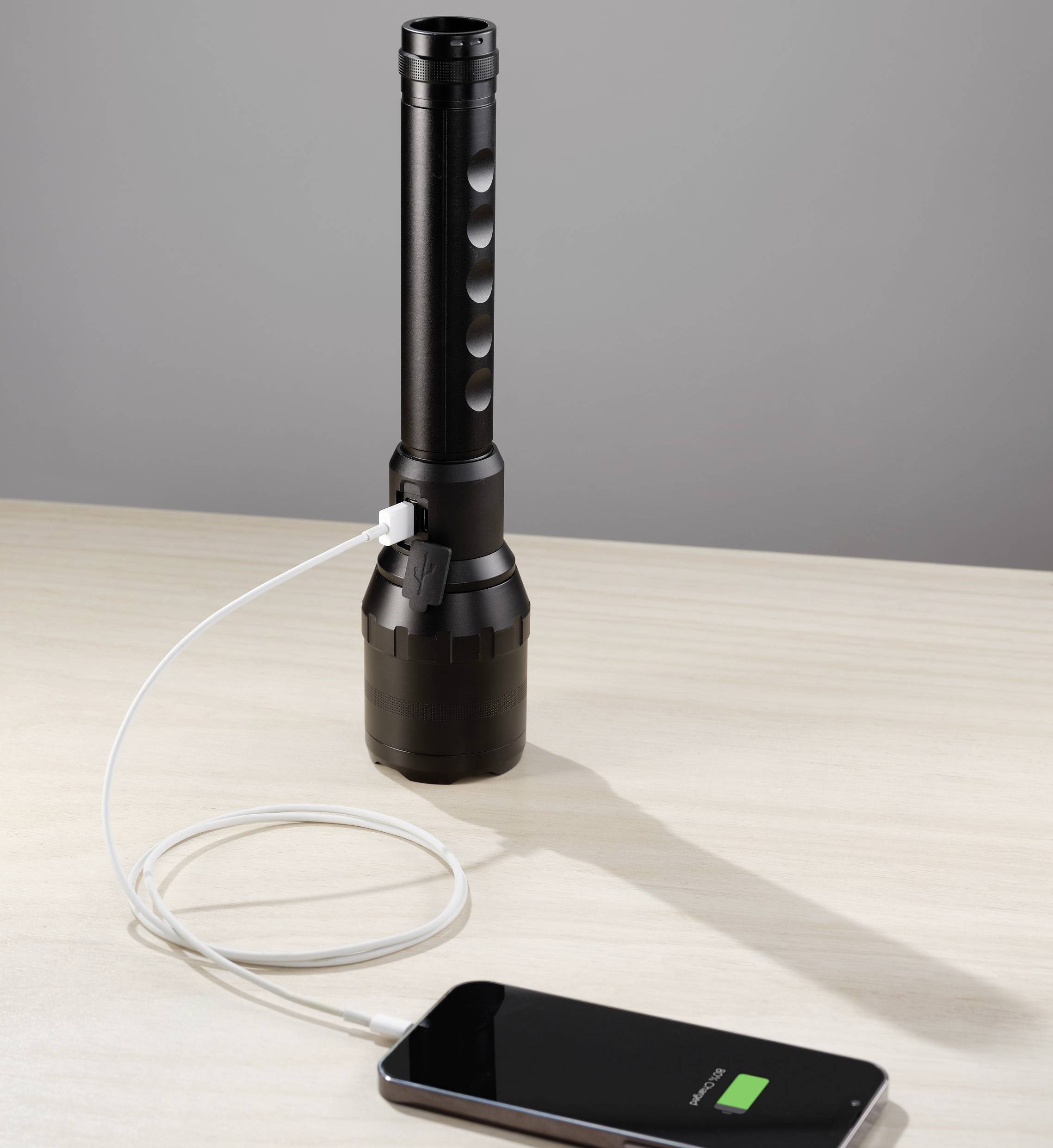 Une lampe de poche charge un smartphone via un câble USB posé sur une table.