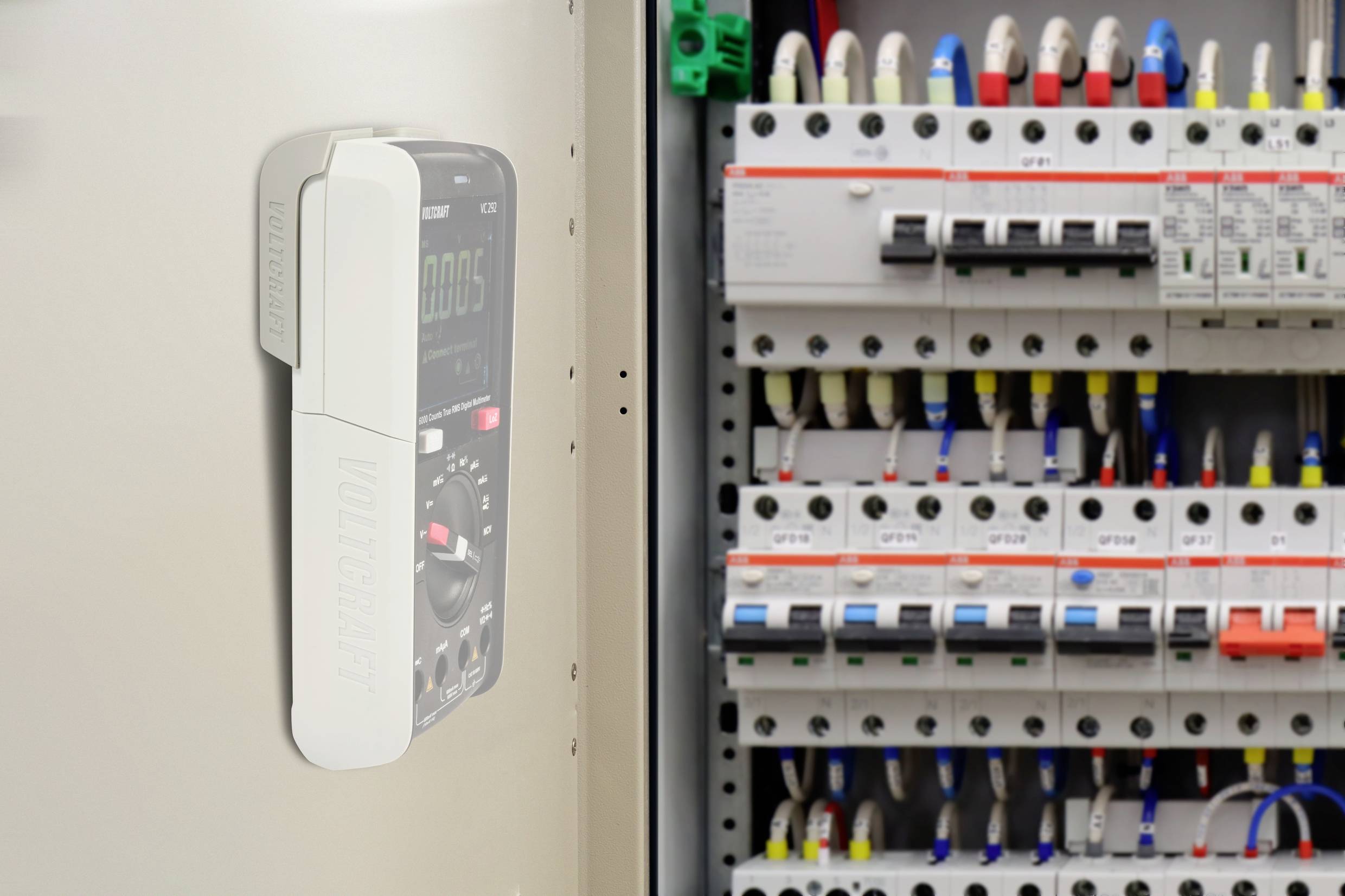 Un multimètre mesure la tension dans un armoire électrique pleine de fusibles et d'interrupteurs. Des panneaux avec différents marquages colorés sont visibles.