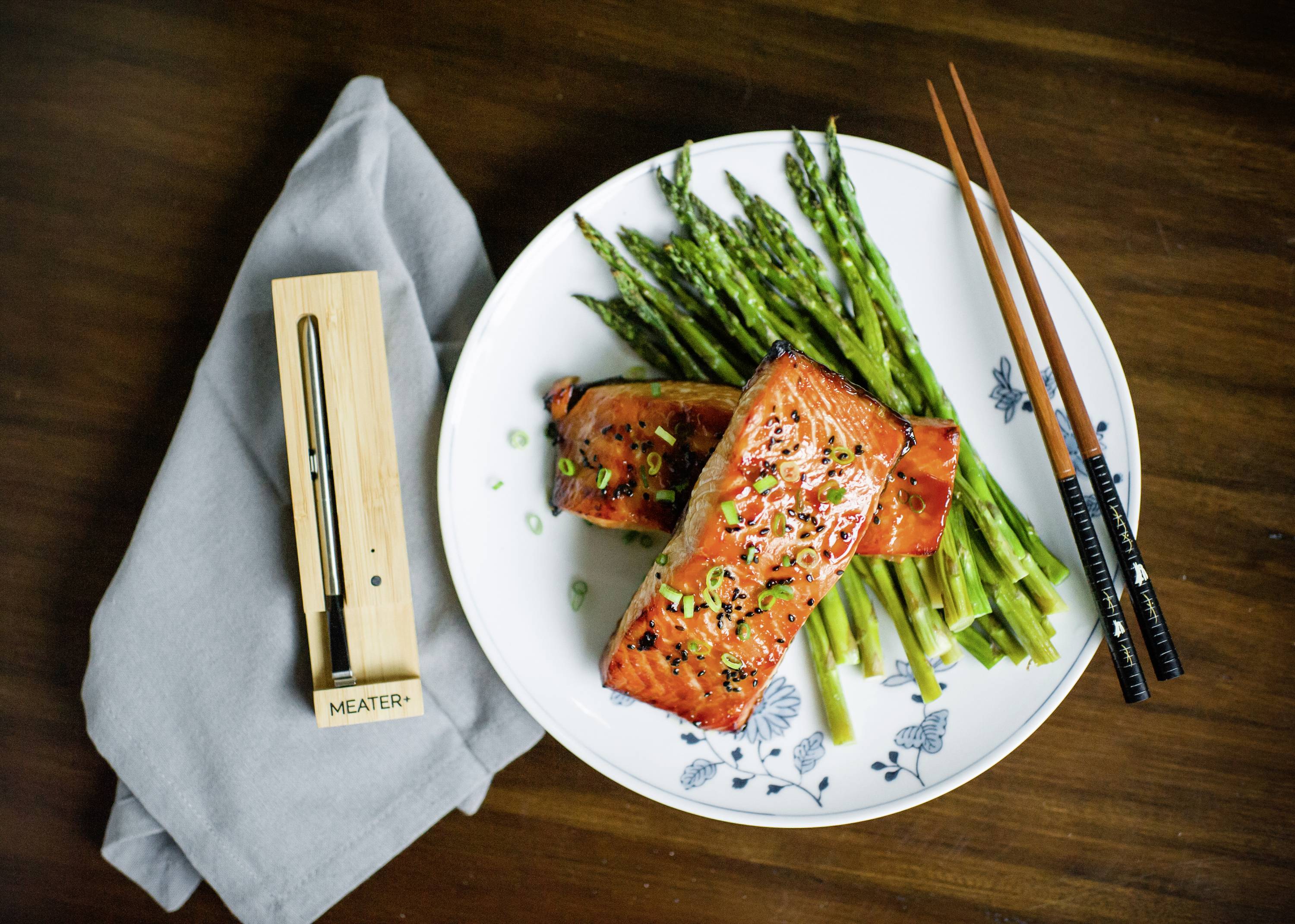 Plat de saumon grillé et d'asperges sur une assiette blanche, à côté un thermomètre de cuisson. Des baguettes sont posées à droite sur la table.