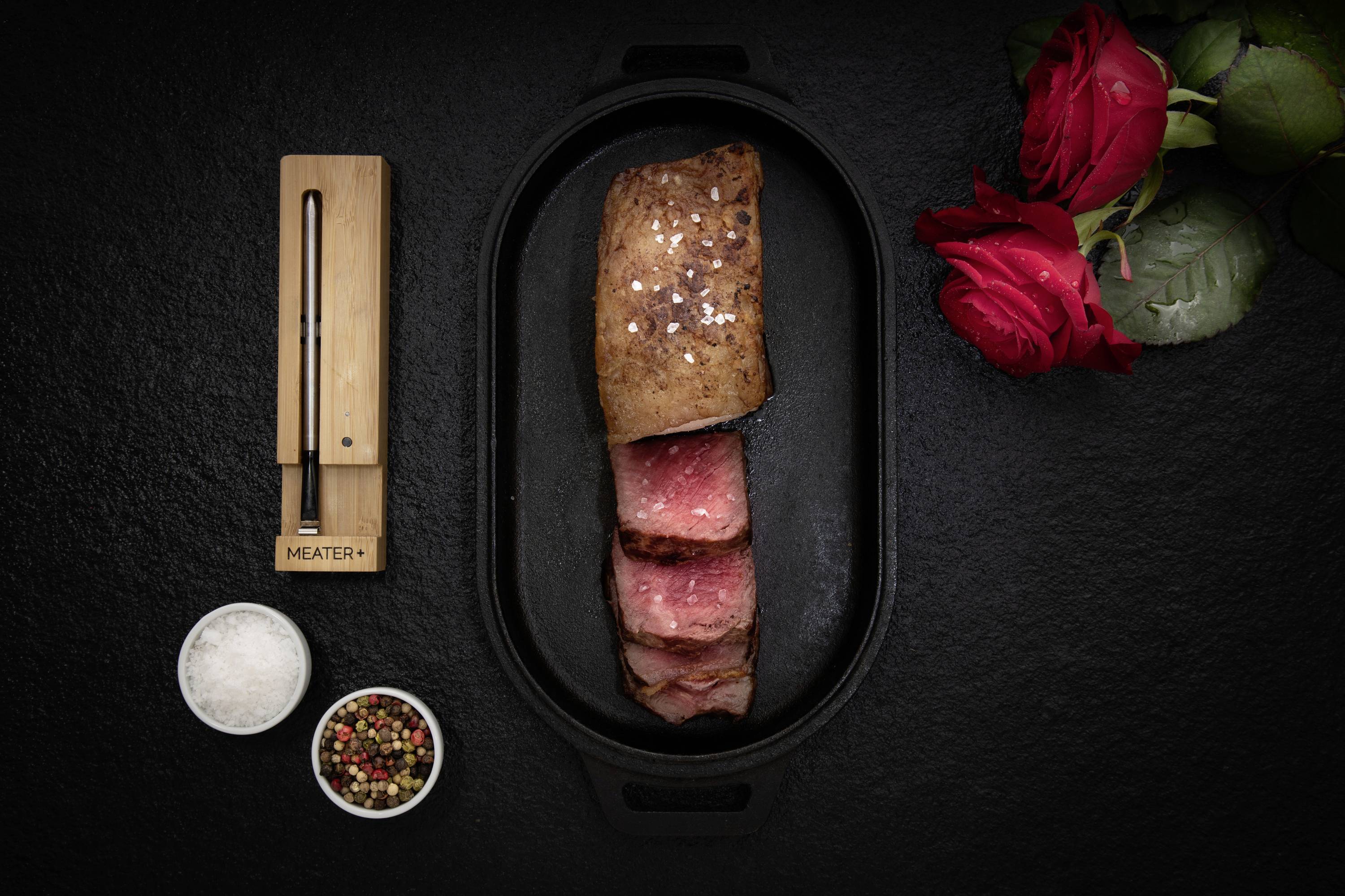 Un steak à point, coupé en deux, dans une poêle noire. À côté, un thermomètre numérique à viande et deux roses rouges.