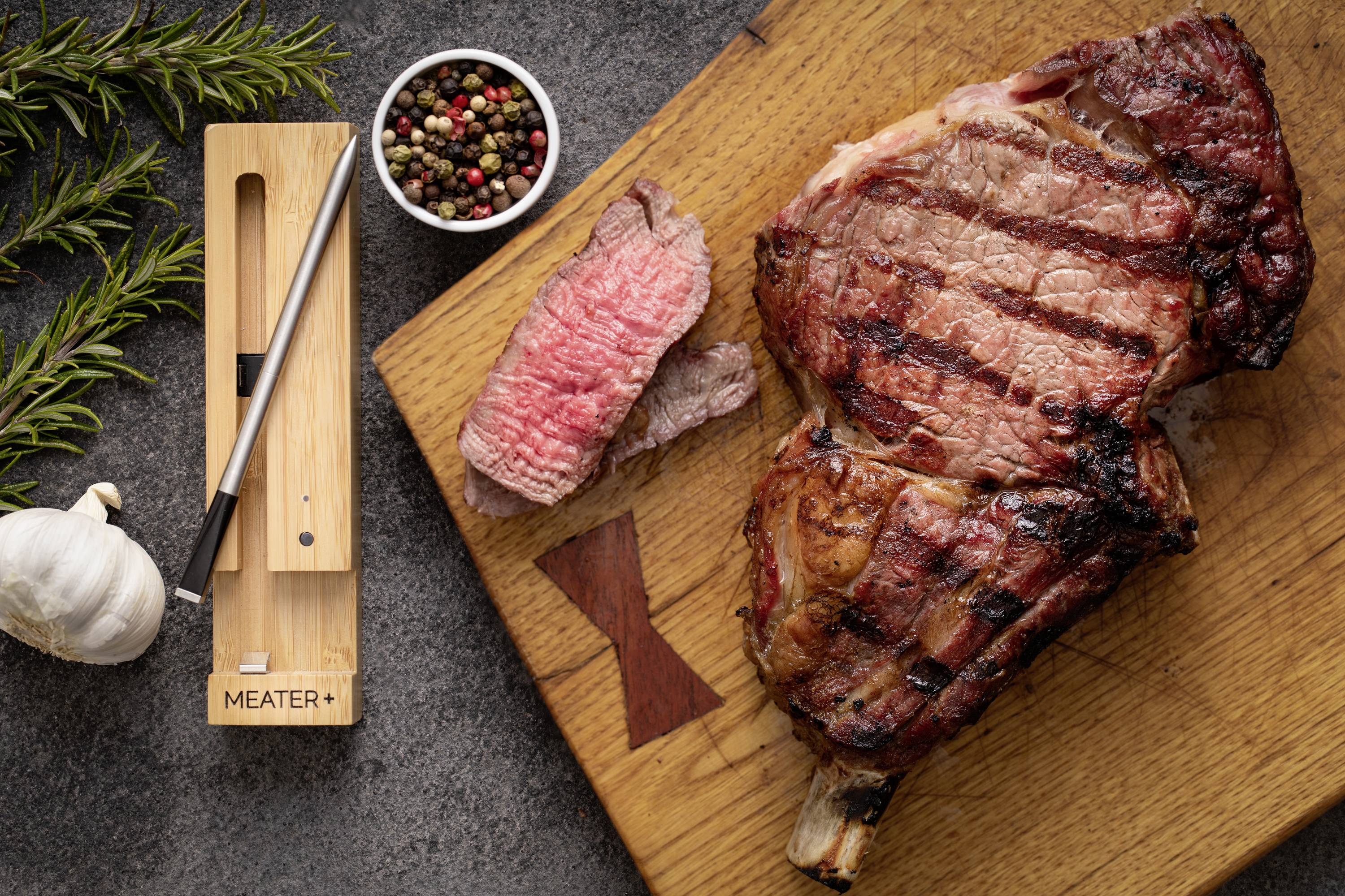 Steak grillé sur une planche en bois, avec un morceau coupé à côté, des grains de poivre, du romarin et de l'ail. Thermomètre à viande à gauche.