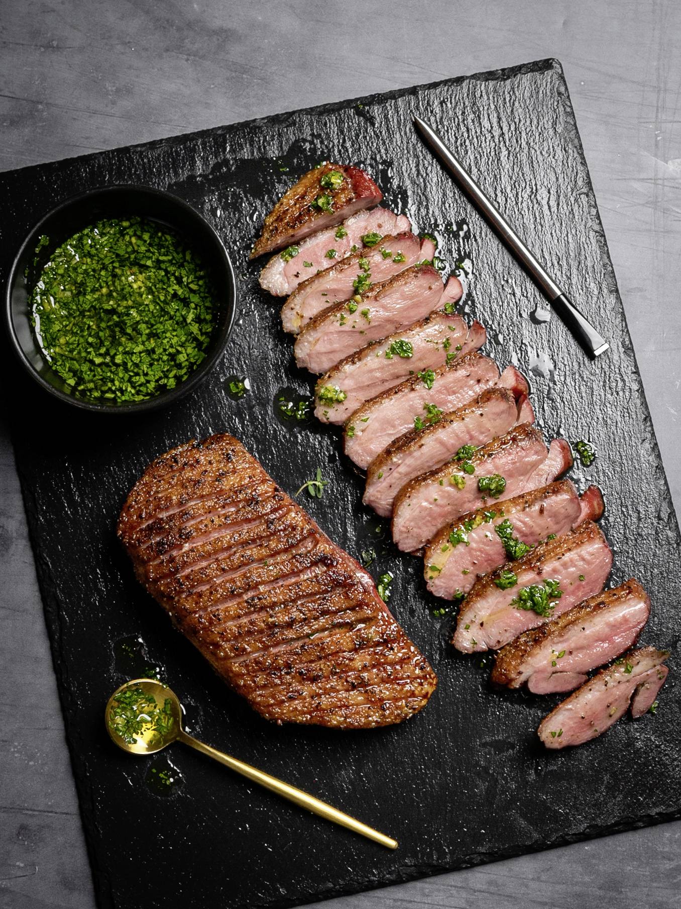 Filet de canard grillé tranché, garni d'huile d'herbes et servi sur une ardoise avec un petit bol de sauce.