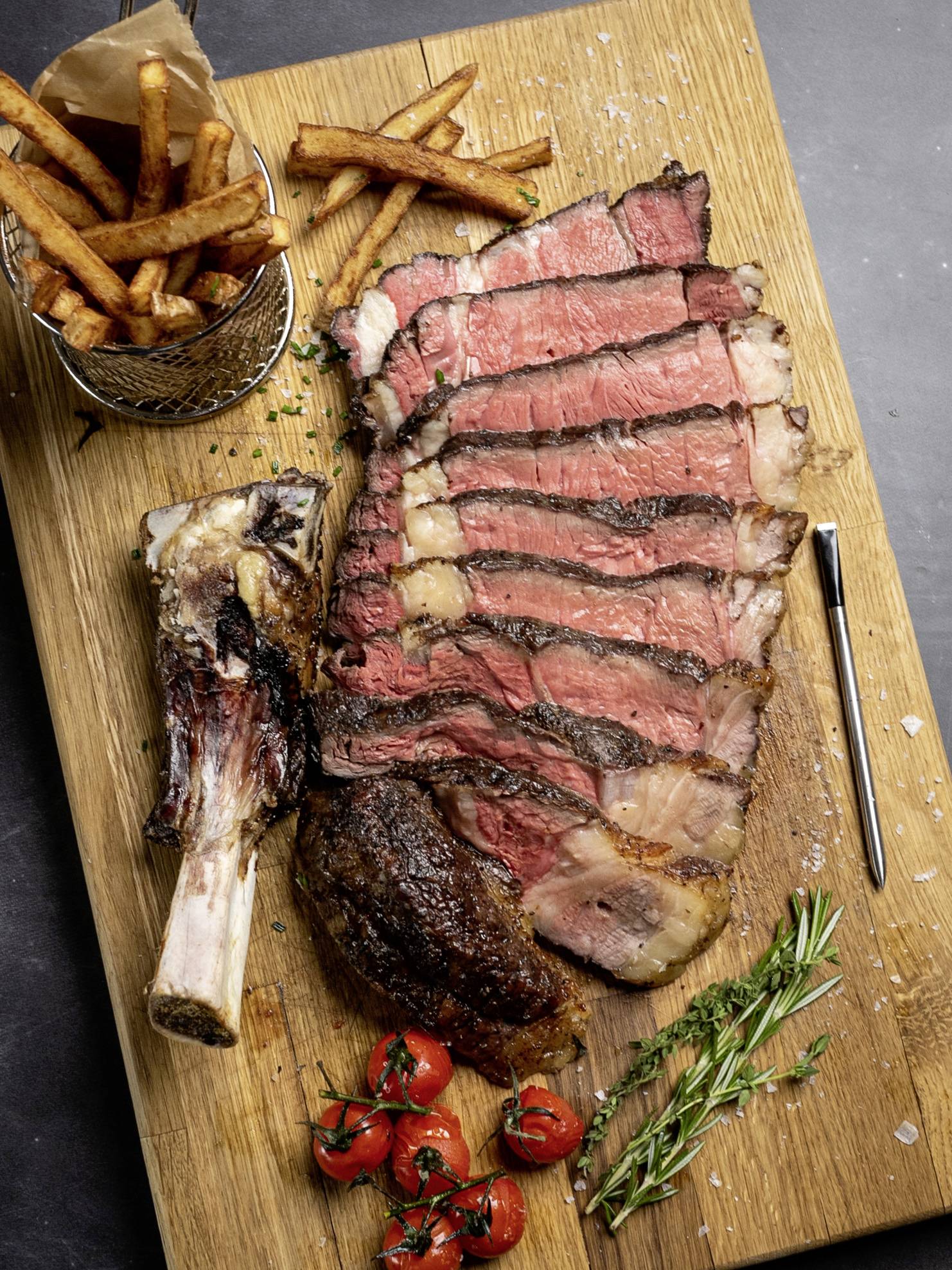 Steak tranché sur une planche en bois avec des frites, des tomates et du romarin.