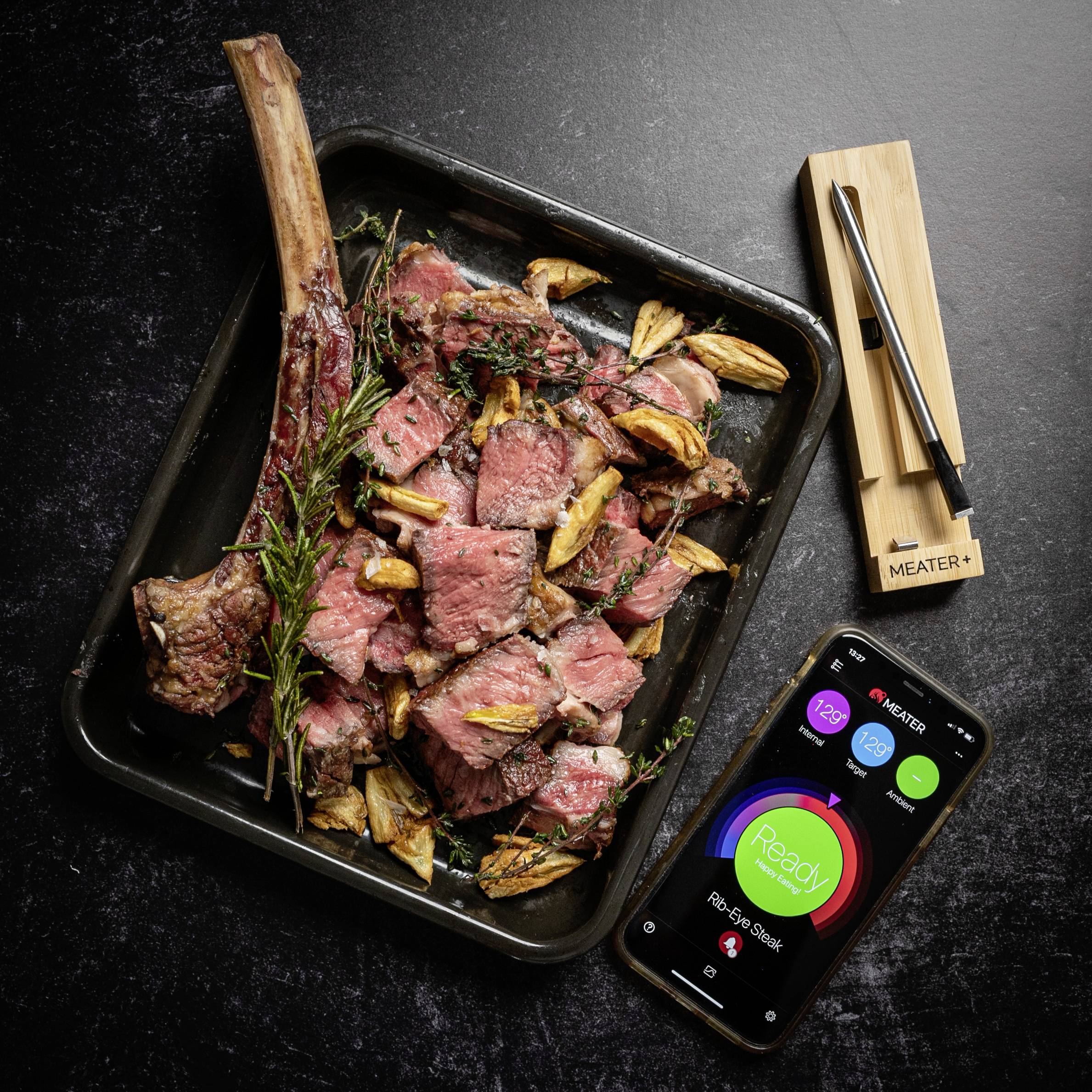 Steak Tomahawk grillé sur un plateau avec de l'ail et du romarin, à côté un thermomètre et un téléphone portable affichant la température.