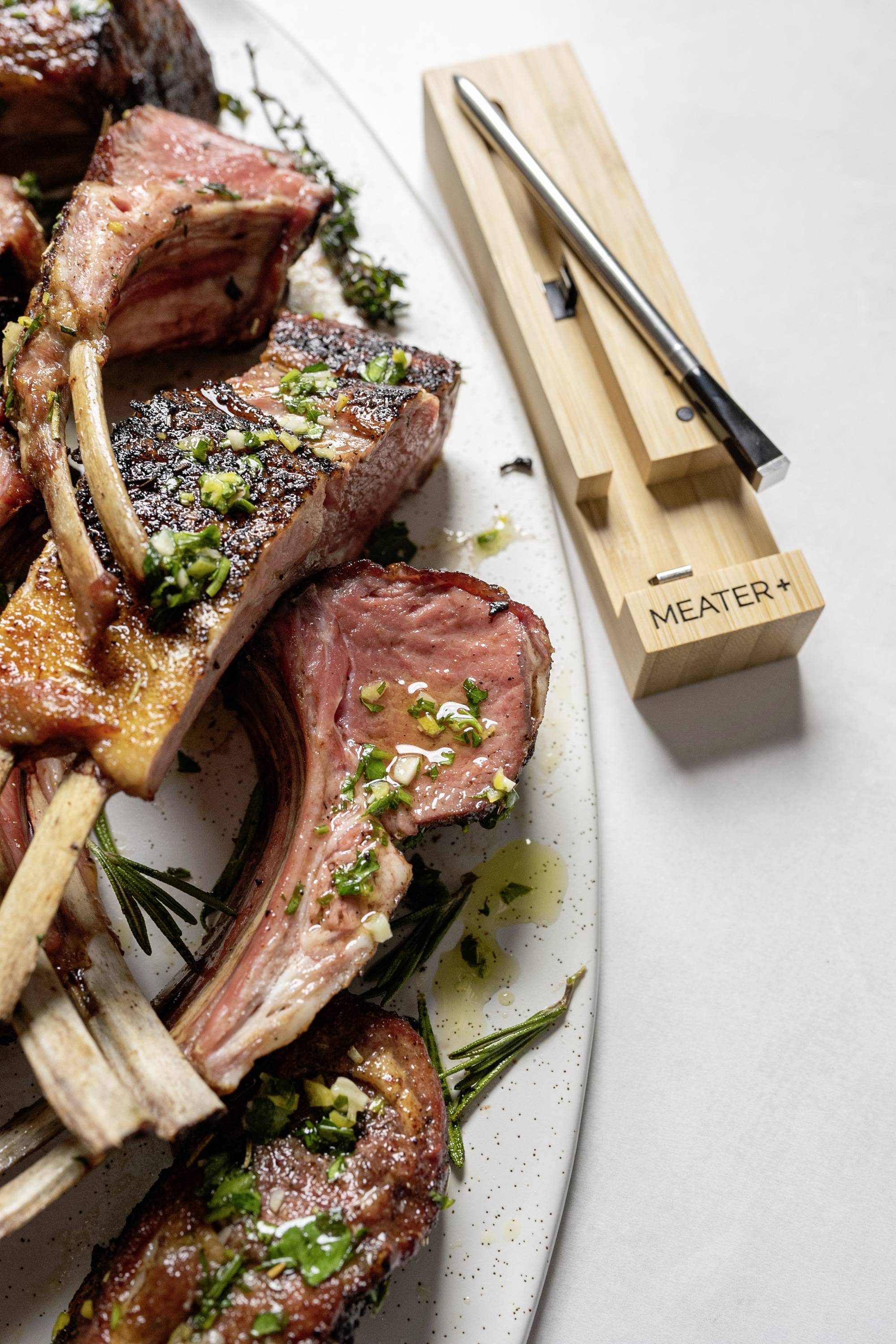 Côtelettes d'agneau servies sur une assiette avec des herbes fraîches, à côté un thermomètre de cuisson posé sur un bloc de bois.