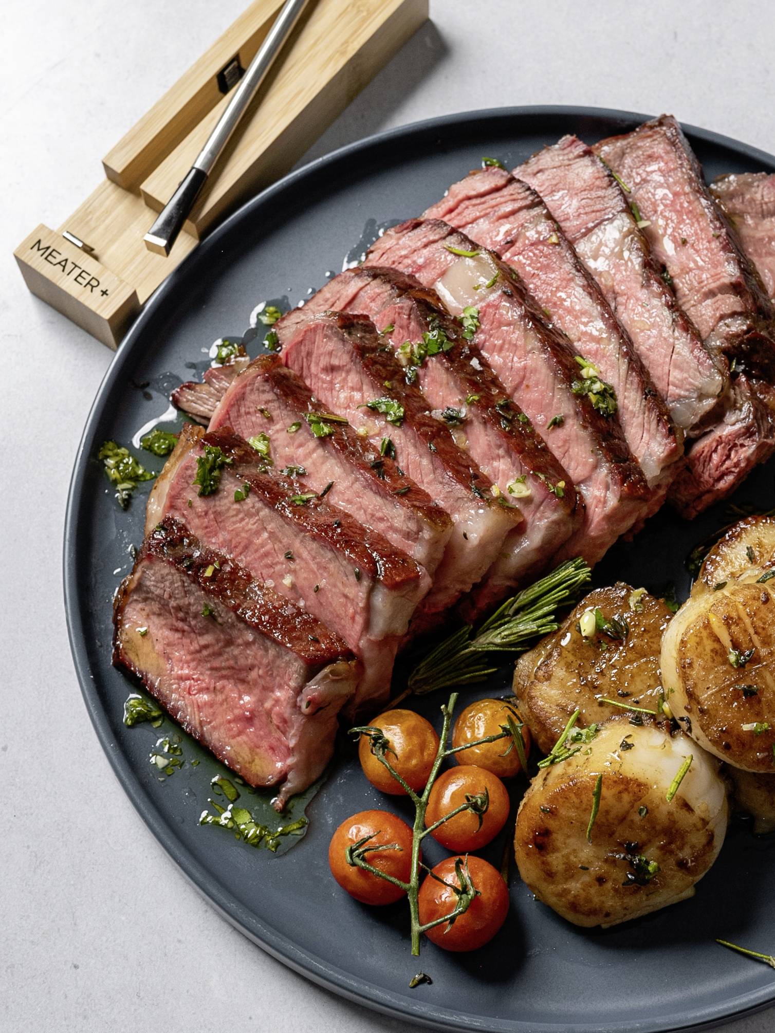 Steak tranché sur une assiette avec des pommes de terre, des tomates cerises et une garniture d'herbes vertes. À côté, deux thermomètres à viande.