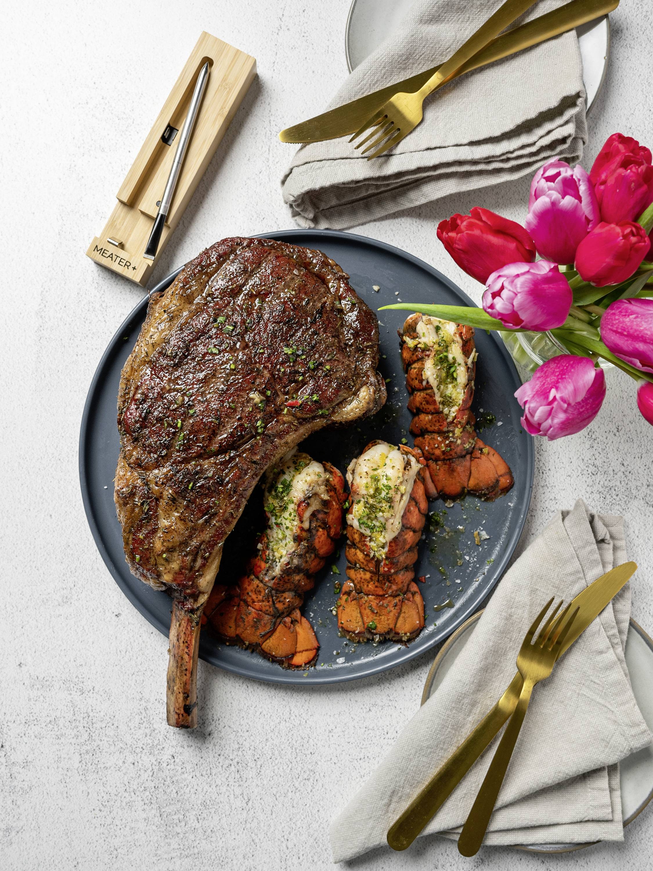 Une assiette garnie d'un steak grillé et de queues de homard, décorée d'herbes fraîches, accompagnée d'un set de couverts et d'un bouquet de tulipes roses et rouges.