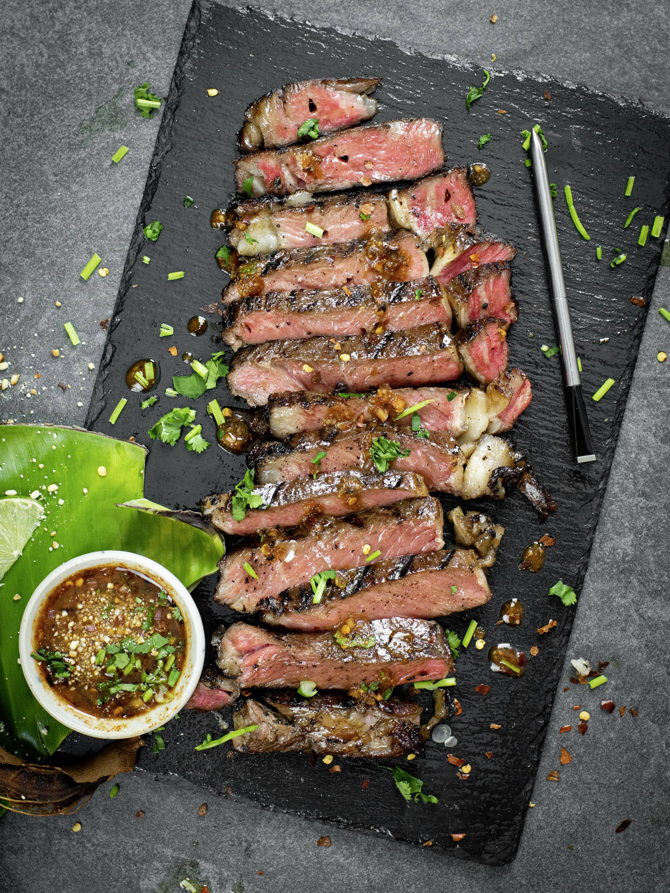 Steak grillé, tranché, servi sur une ardoise noire avec une tasse de sauce et des herbes fraîches en garniture.