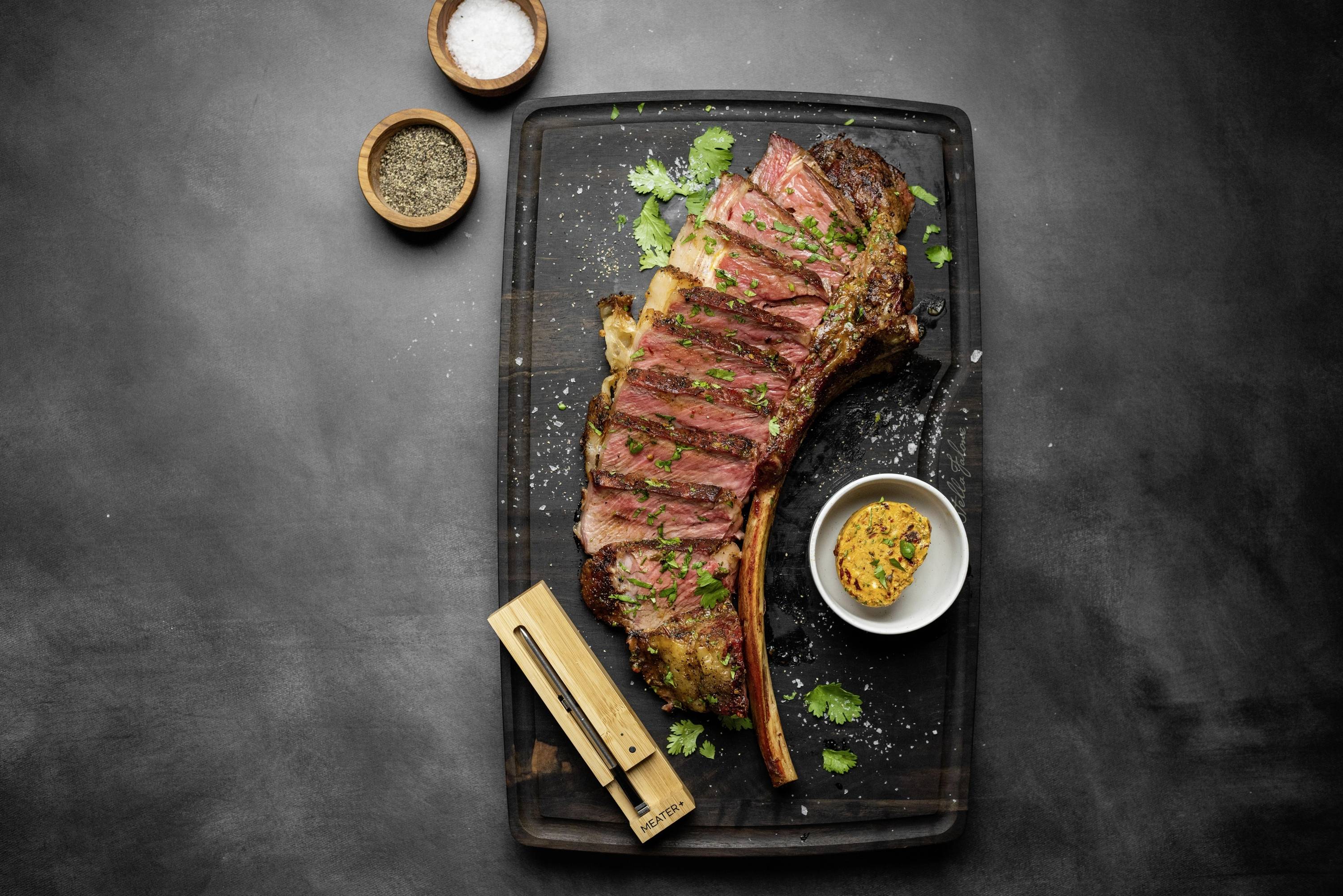 Un steak Tomahawk grillé sur une planche, coupé en tranches, garni d'herbes, avec du sel, du poivre et du beurre aux herbes à côté.