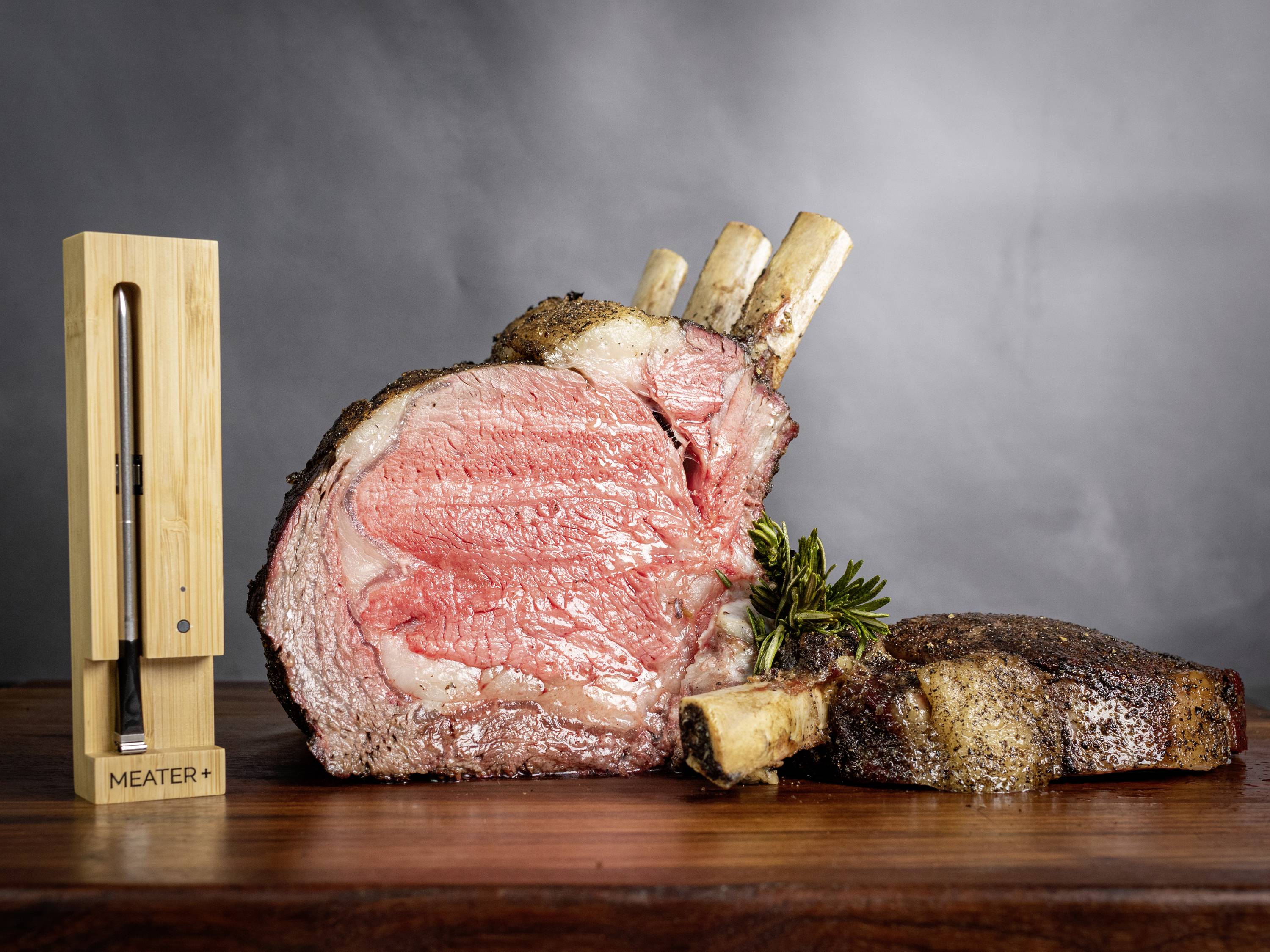 Un grand morceau de bœuf juteux avec os est posé sur une planche à découper. À côté se trouve un thermomètre à viande dans son support en bois.