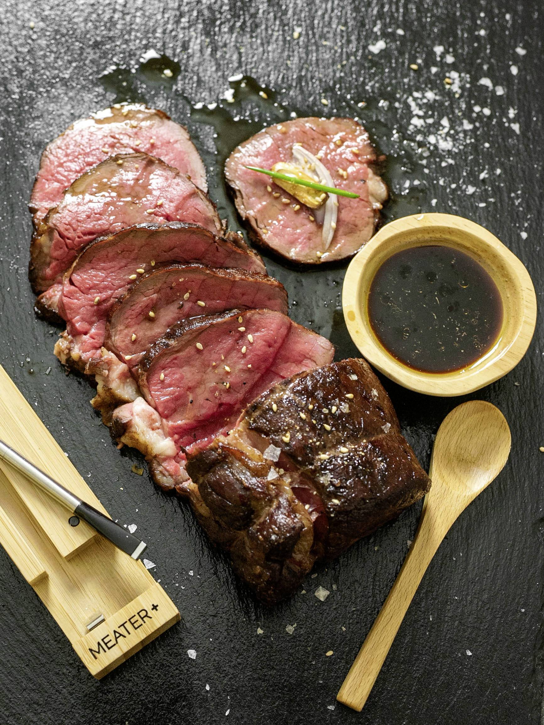Rosbif tranché, disposé sur une ardoise noire, accompagné d'une sauce dans un bol et d'une cuillère en bois à côté.
