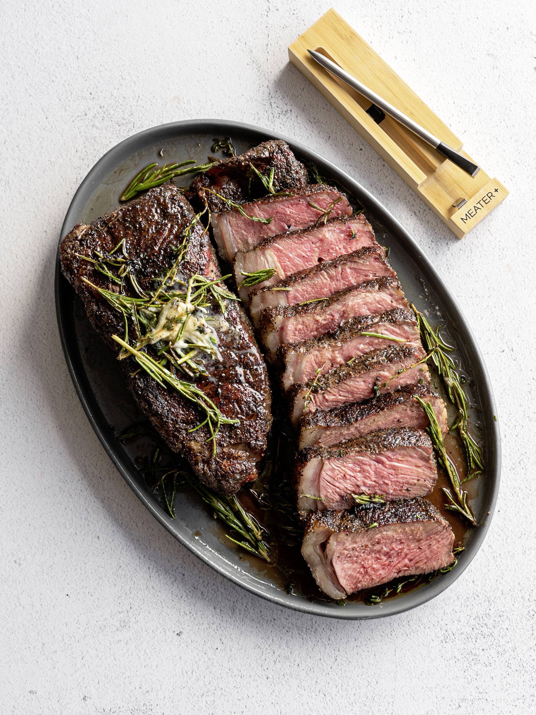 Sur une assiette ovale noire sont disposés deux morceaux de steak juteux et bien dorés, l'un entier et l'autre tranché, garnis de romarin.