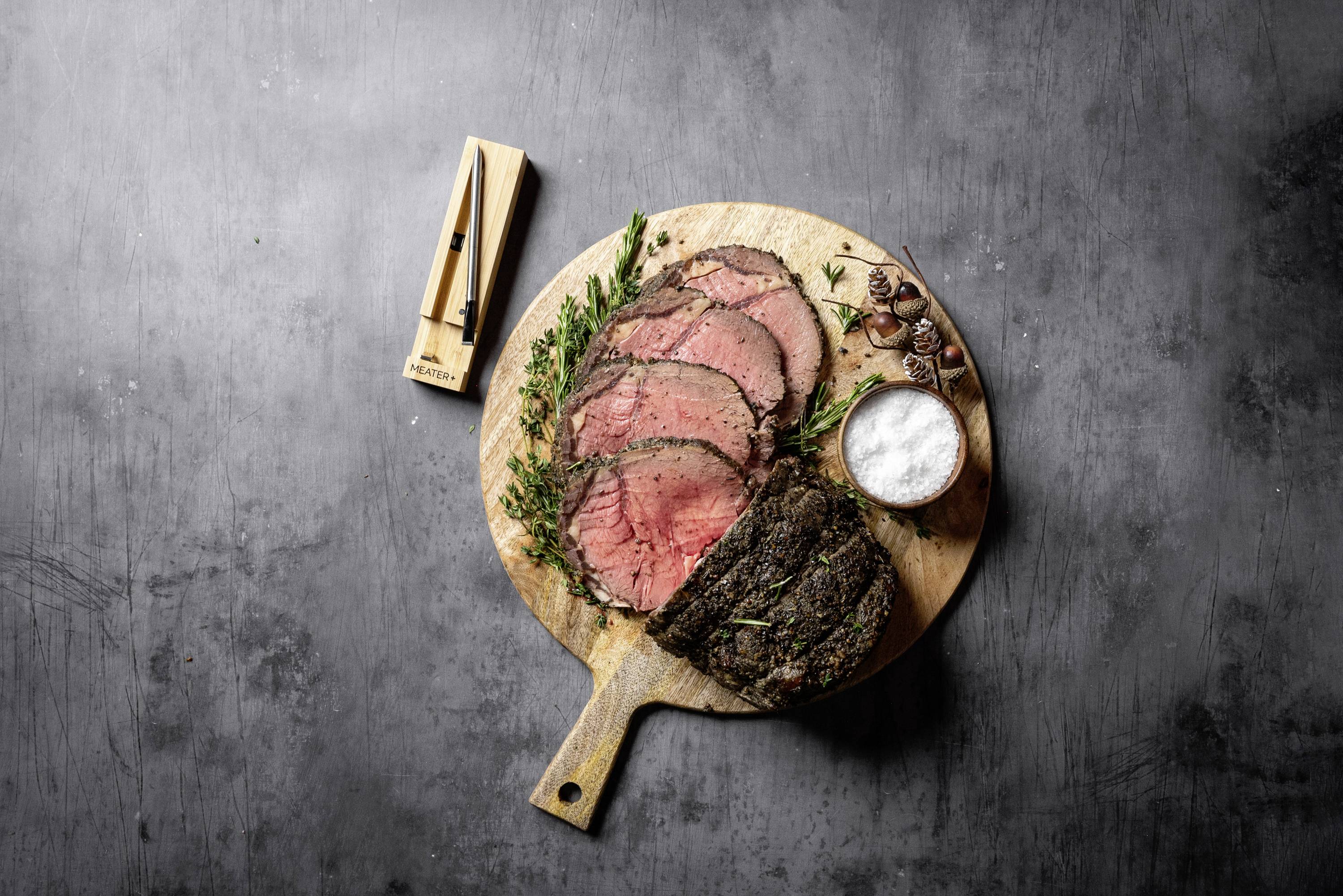 Rosbif tranché sur une planche en bois, garni d'herbes, accompagné de sel et d'un couteau. Idéal pour un dîner élégant.
