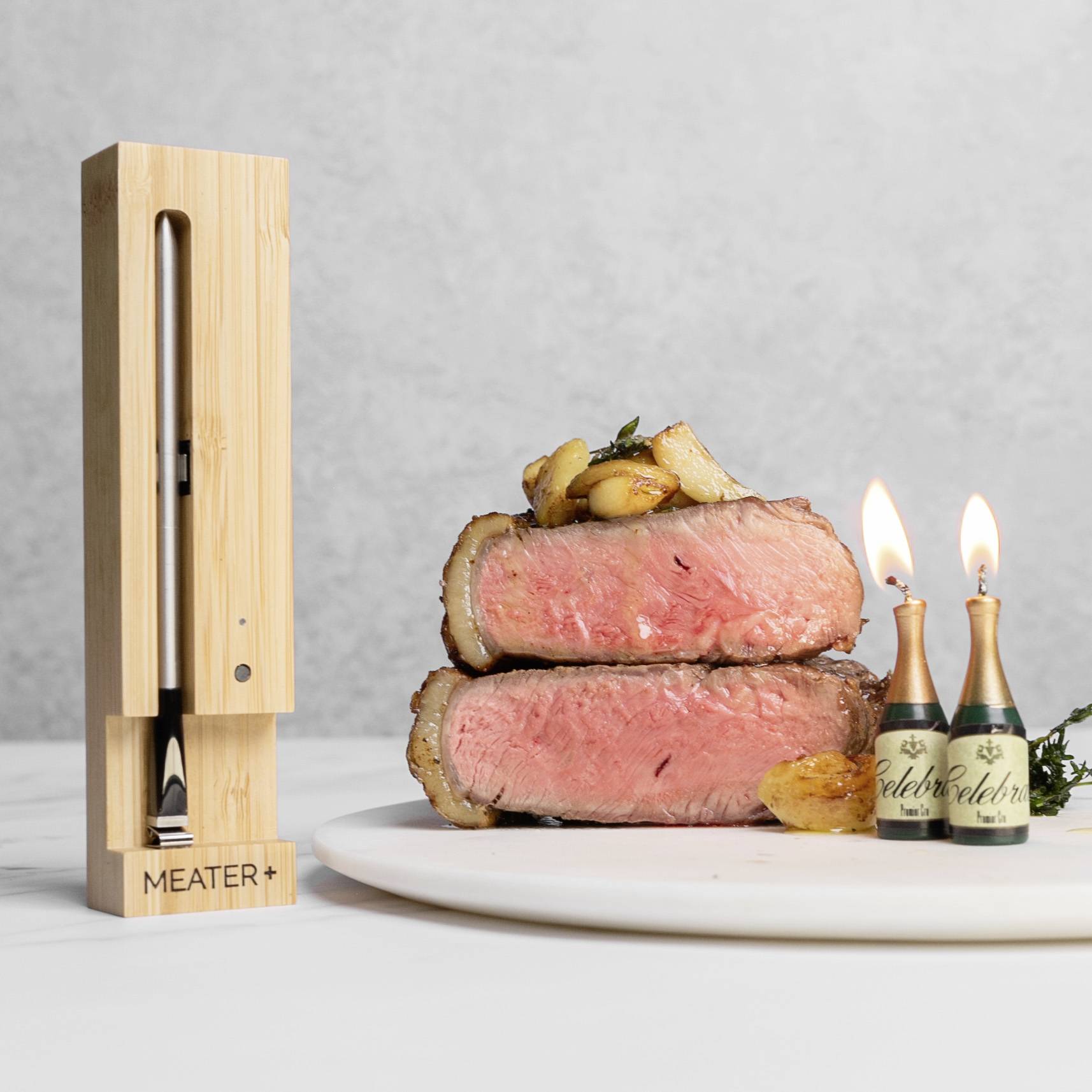 Une pile de viande cuite saignante sur une assiette. À côté, deux bougies allumées. À gauche, un thermomètre en bois avec l'inscription 'MEATER+'.
