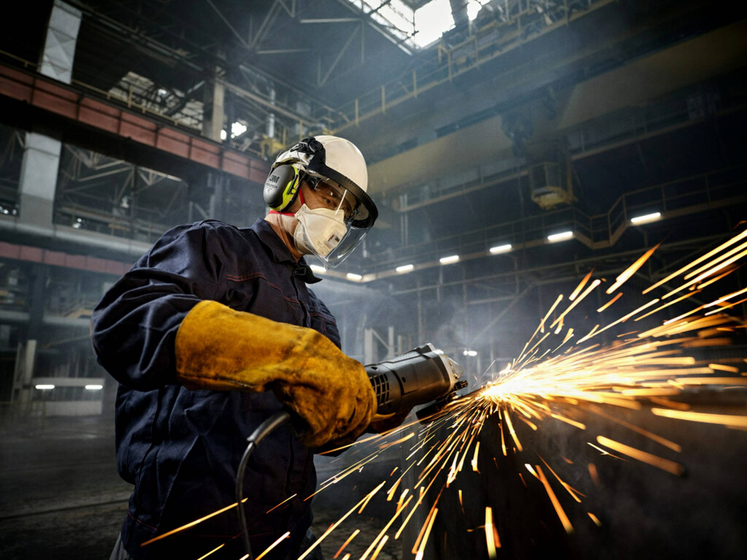 Un ouvrier en tenue de protection meule du métal dans un hall d'usine. Des étincelles jaillissent de la meuleuse.