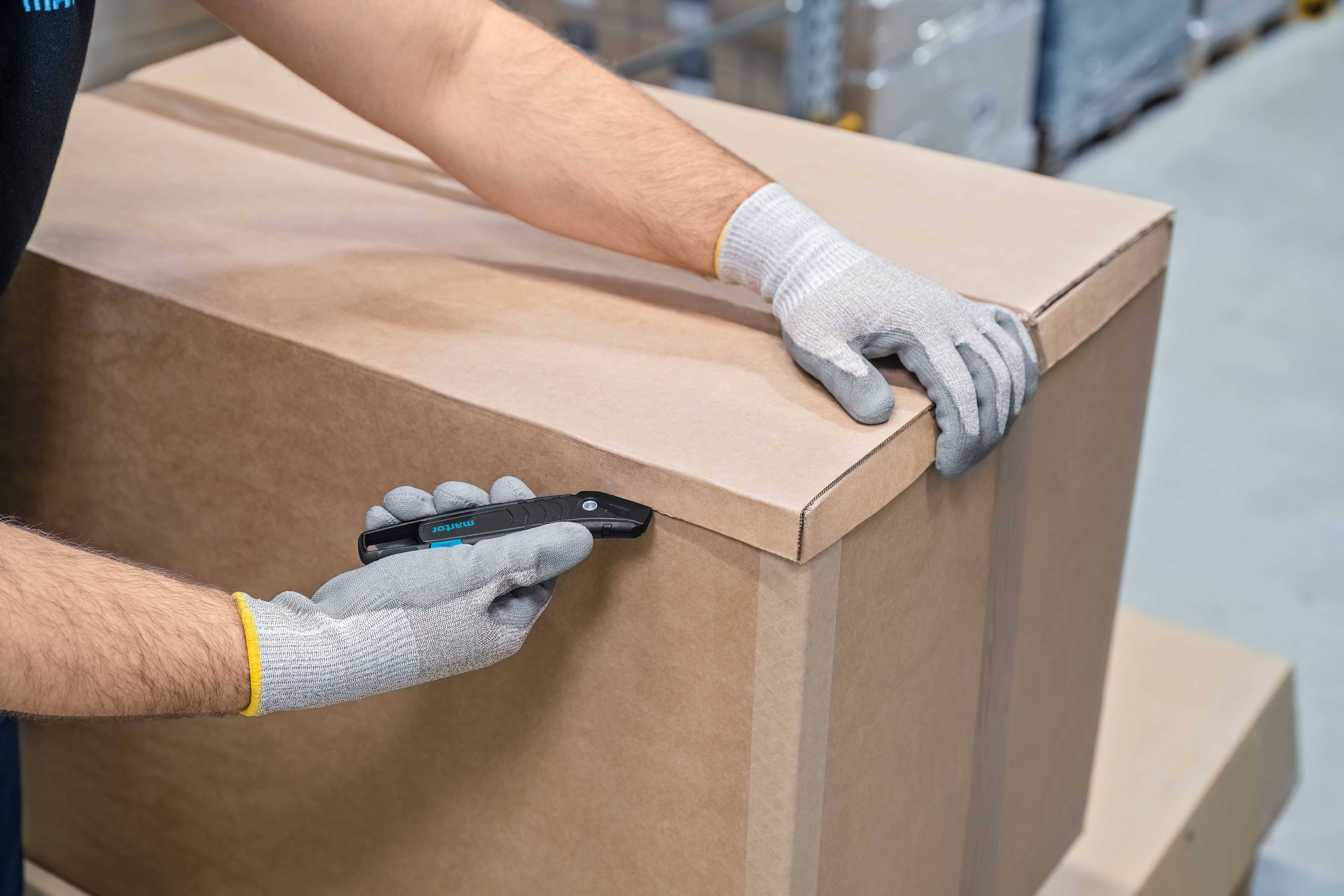 Une personne portant des gants gris ouvre prudemment un grand carton à l'aide d'un cutter noir dans un environnement d'entrepôt.