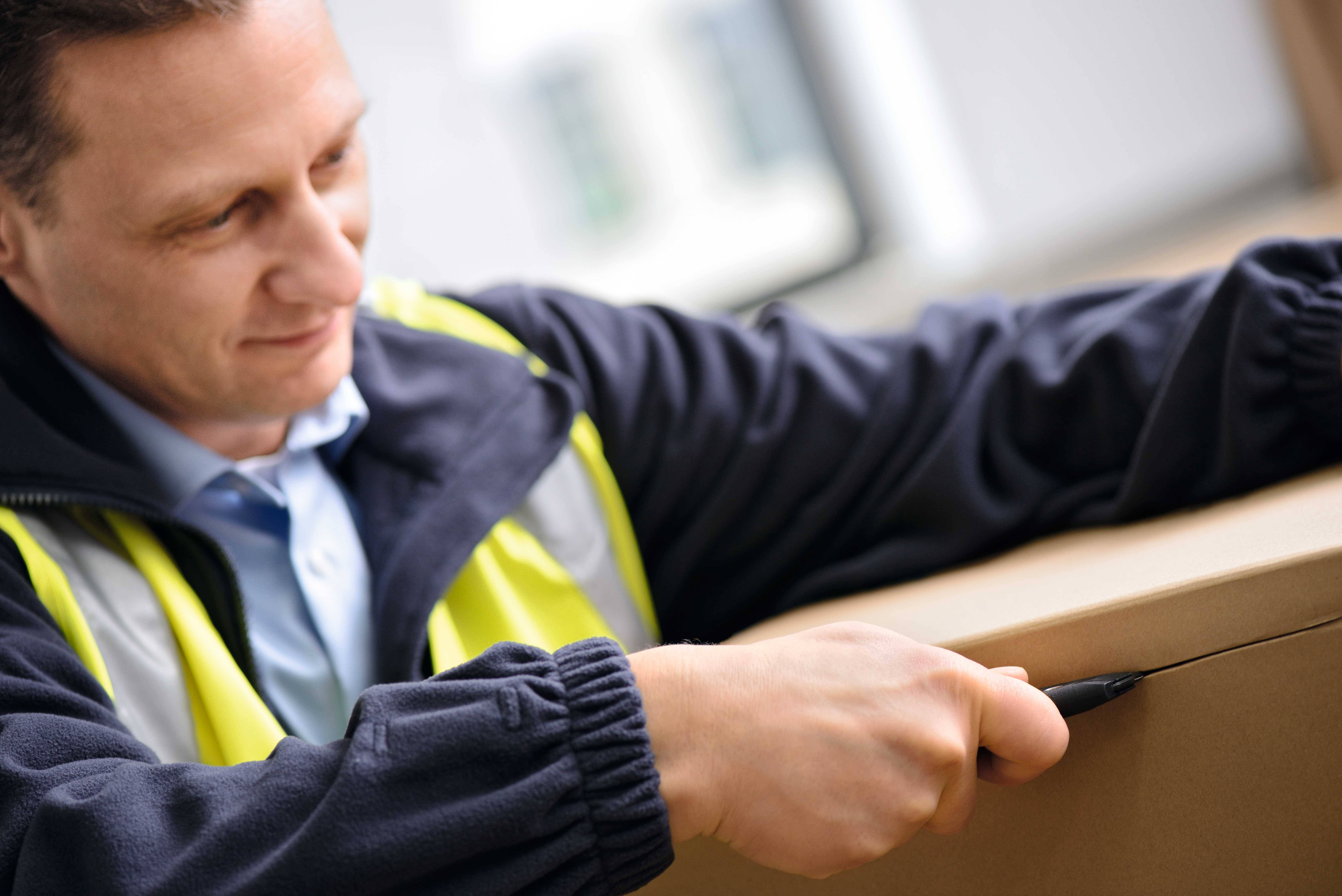 Un homme en tenue de travail ouvre un carton à l'aide d'un cutter. Il porte un gilet de sécurité et semble concentré sur sa tâche.
