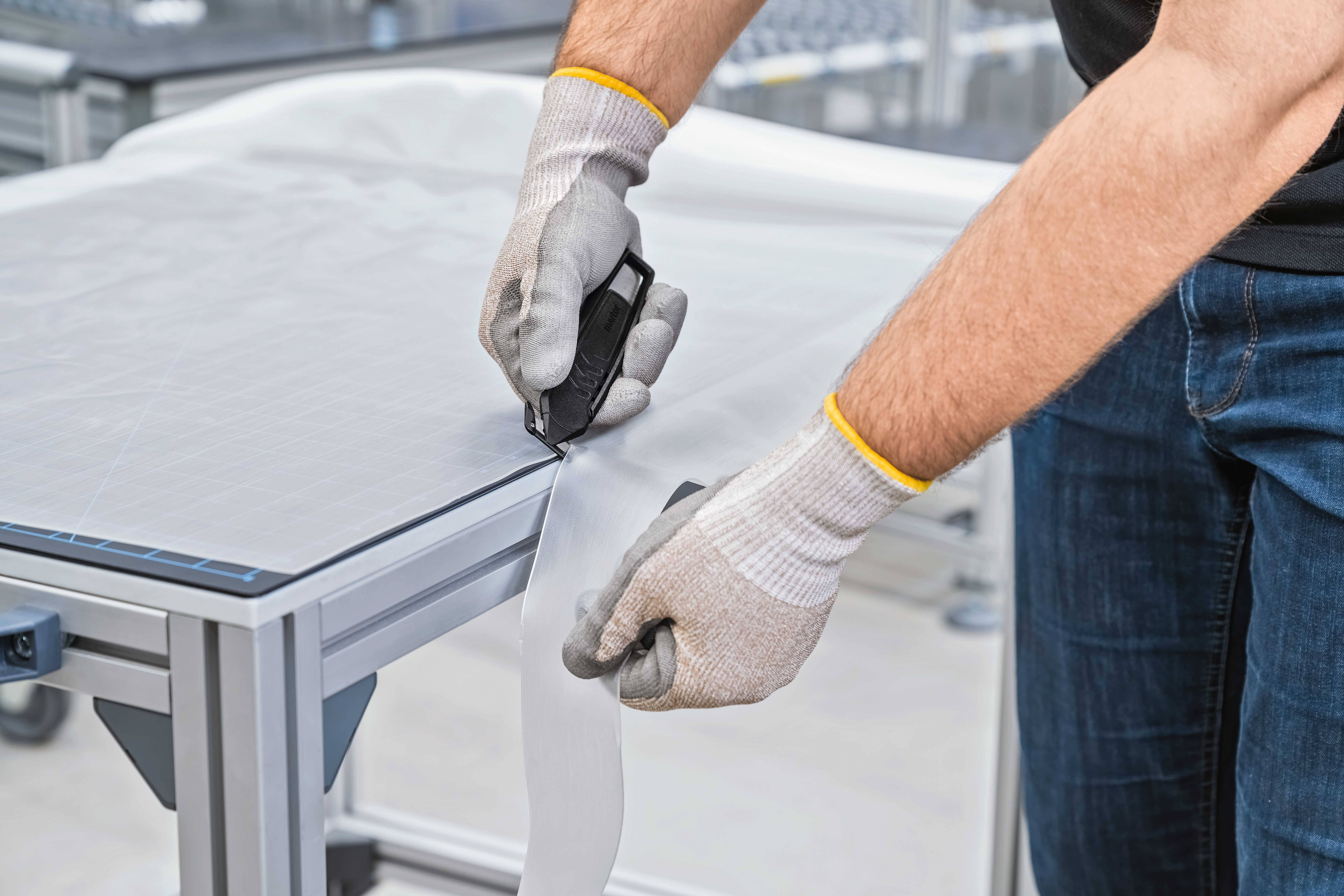 Une personne portant des gants coupe un matériau blanc sur une table à l'aide d'un cutter.
