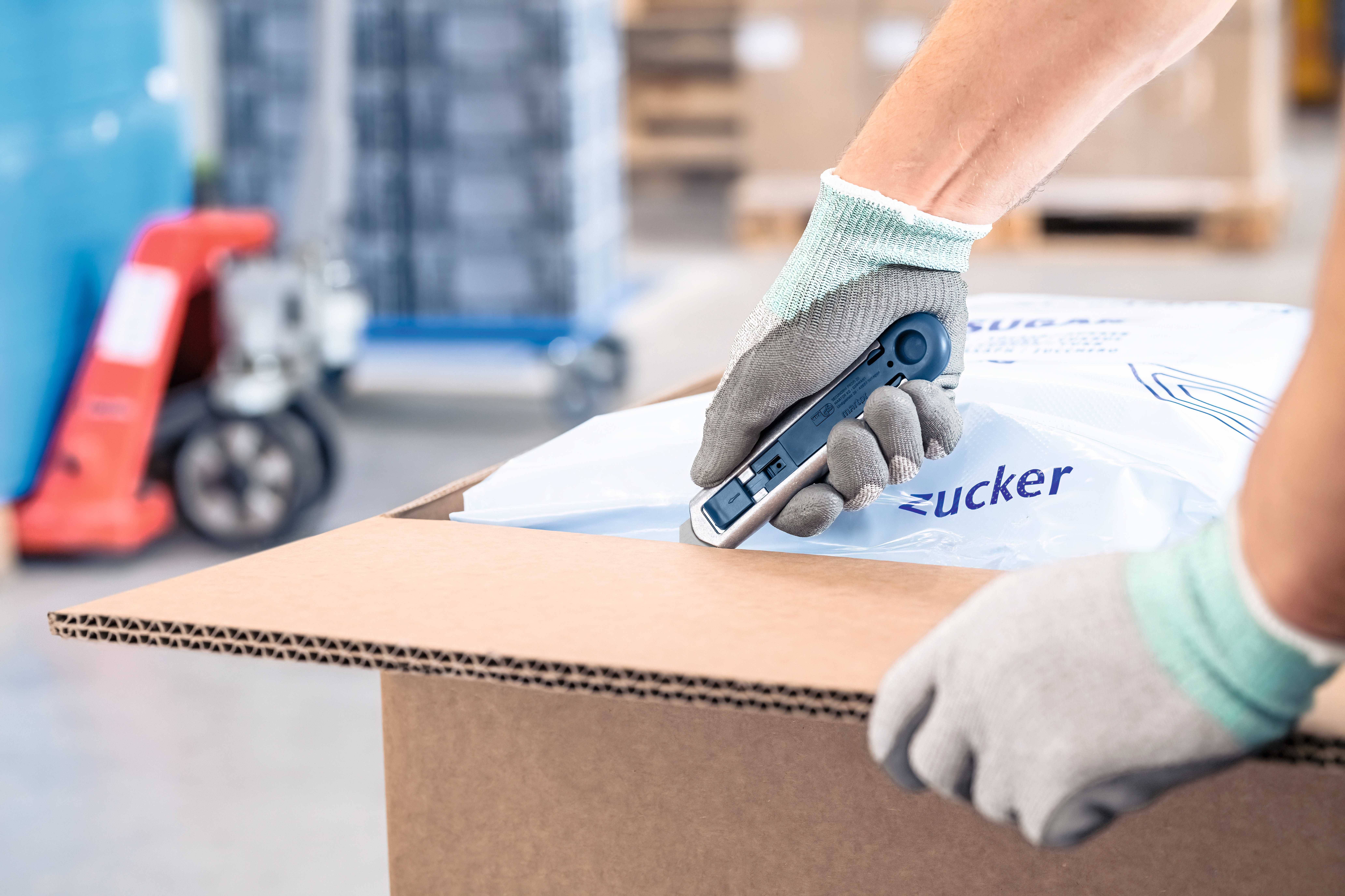Deux mains gantées ouvrent un carton à l'aide d'un cutter. À l'arrière-plan, des palettes et des emballages sont visibles de manière floue.