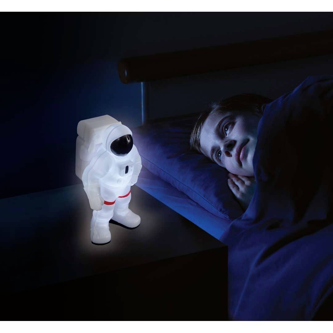 Un enfant est couché dans son lit et regarde une lampe d'astronaute lumineuse posée sur la table de chevet. La lampe éclaire doucement la pièce.
