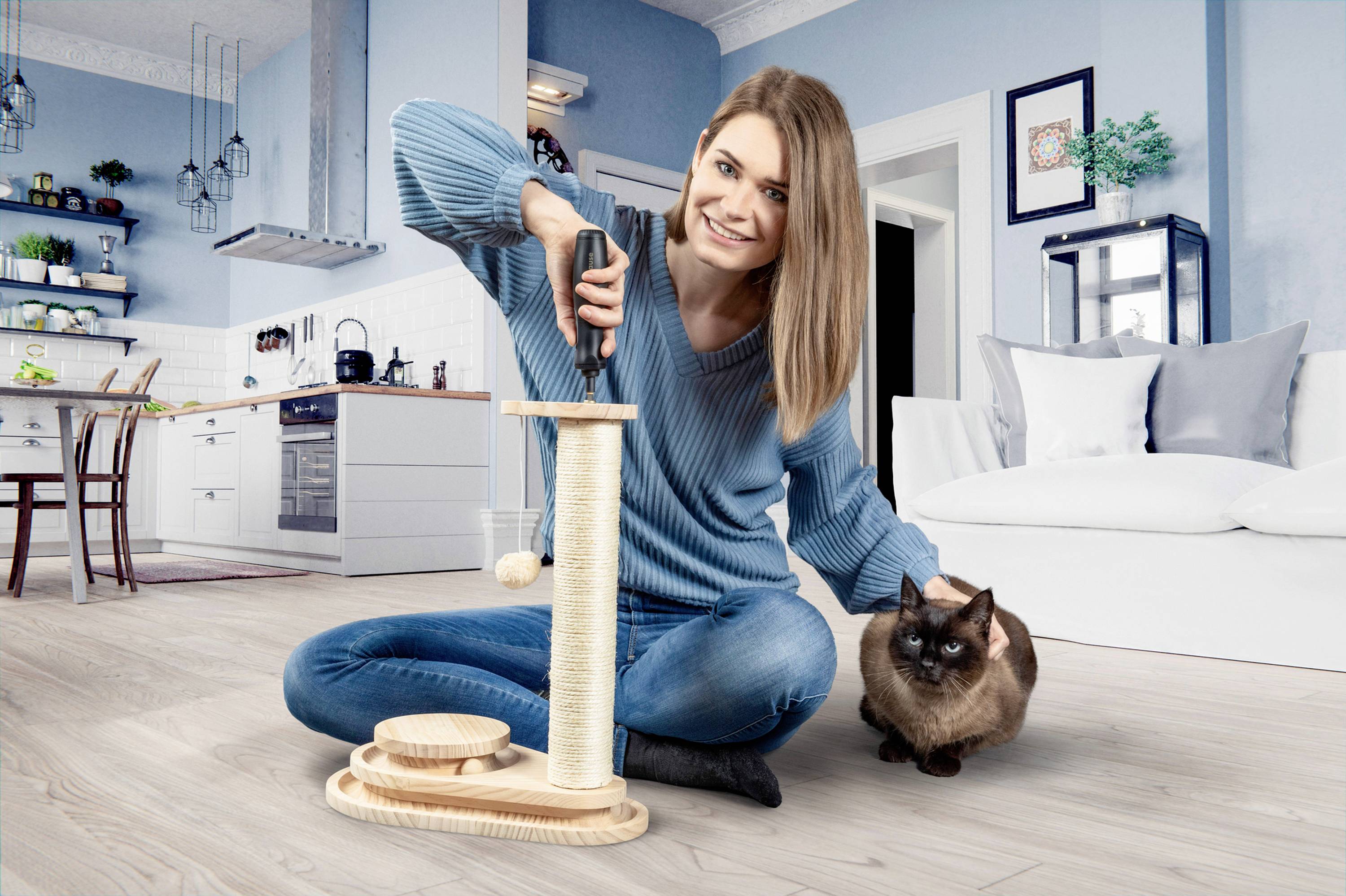 Une femme portant un pull bleu est assise par terre et joue avec un chat. Elle utilise un jouet pour chat dans le salon.