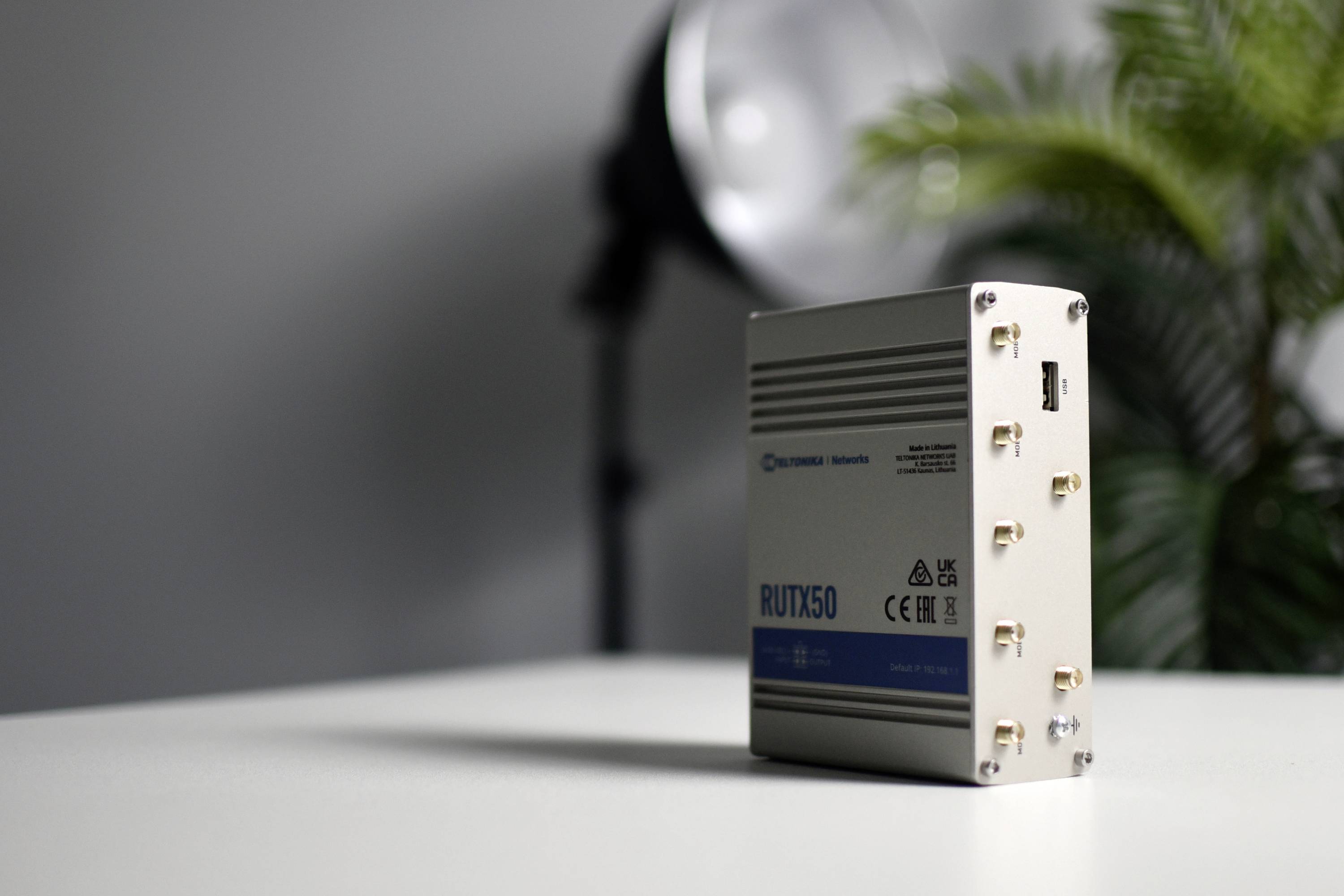 Petit appareil électronique gris argenté portant l'inscription 'RUTX50' posé sur une table, avec une plante et une lampe en arrière-plan.