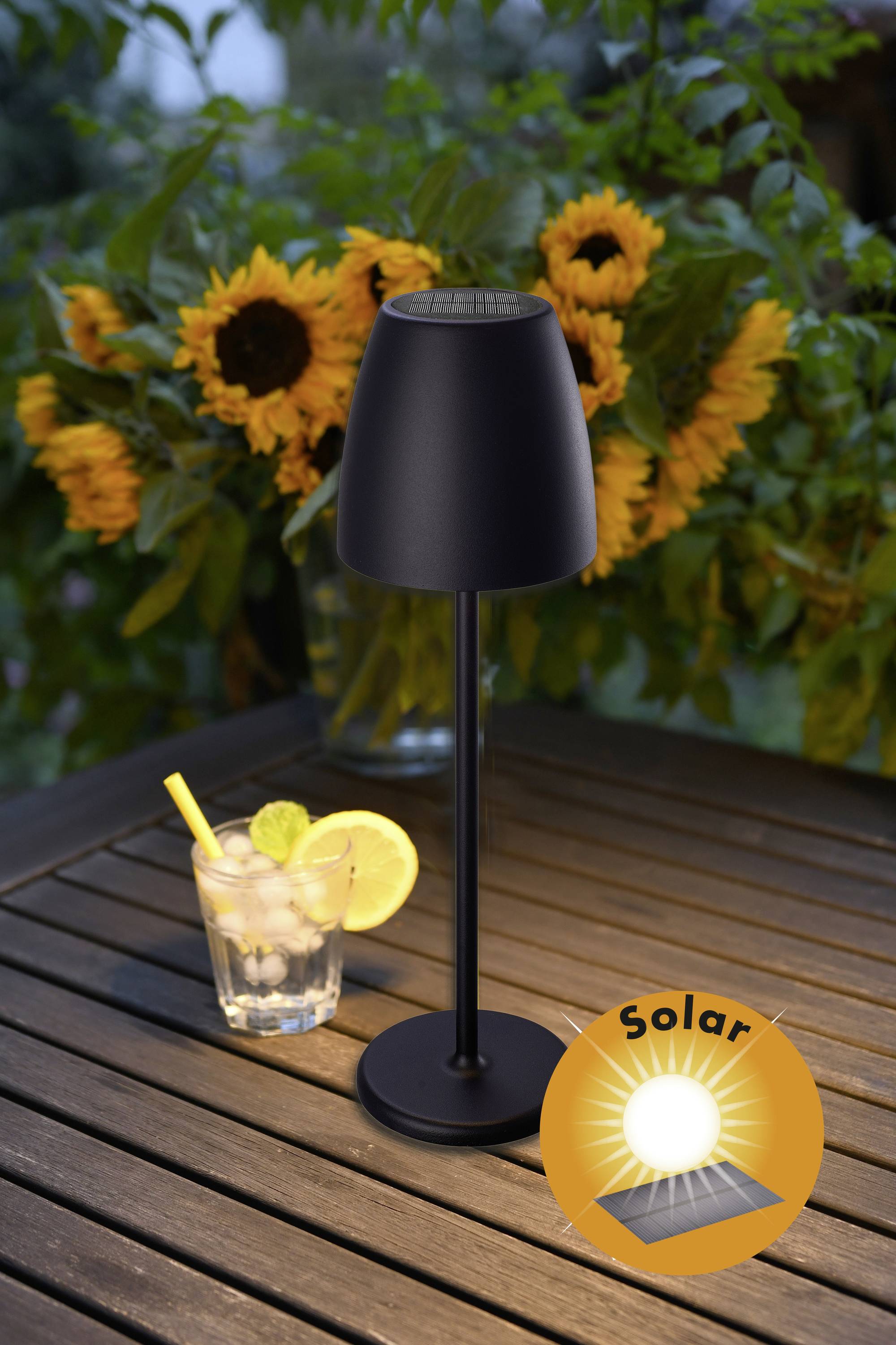 Une lampe solaire noire est posée sur une table en bois devant un bouquet de tournesols. À côté, un verre d'eau glacée avec un citron.