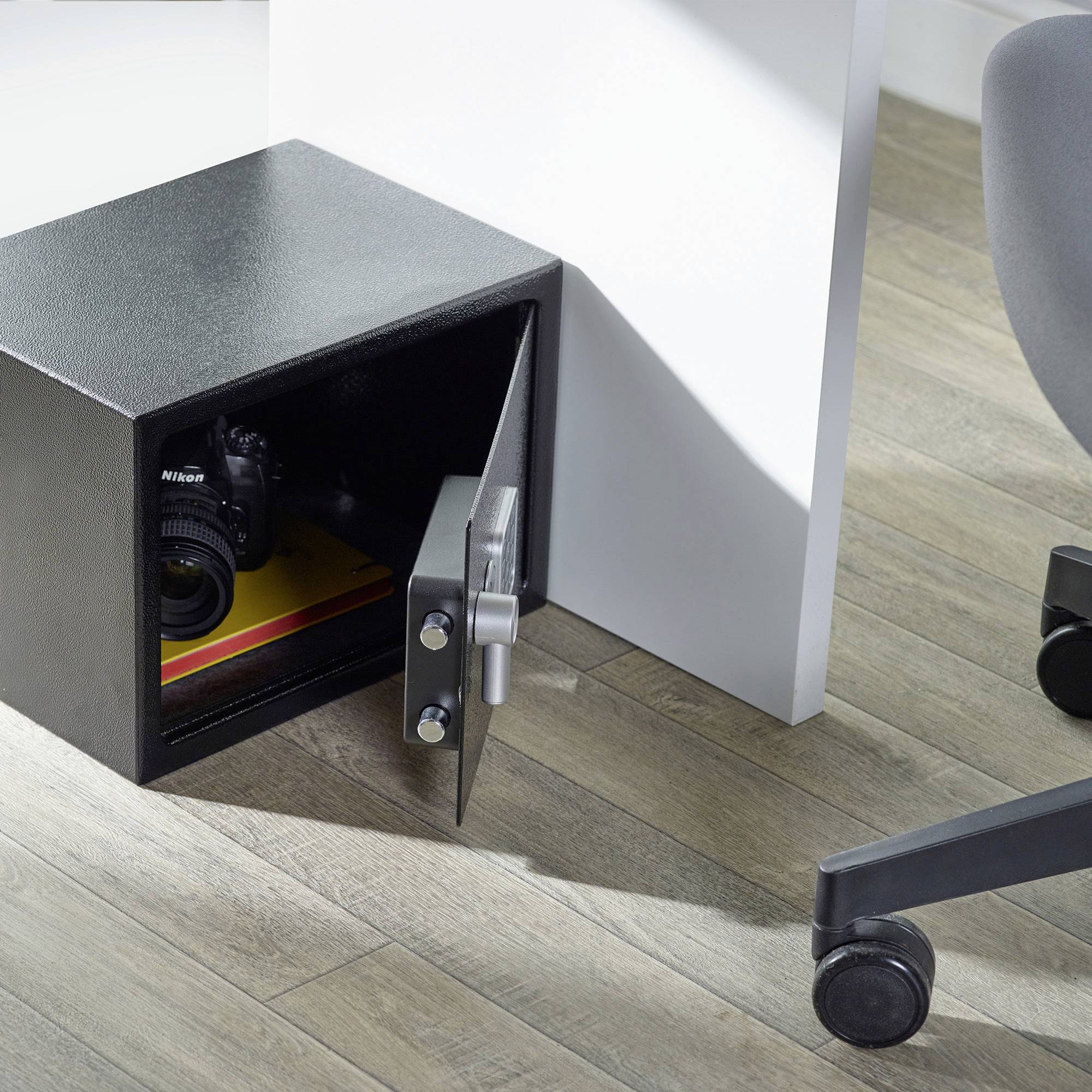 Un coffre-fort noir ouvert sur un plancher en bois contient un appareil photo et un livre. À côté se trouve une chaise de bureau.