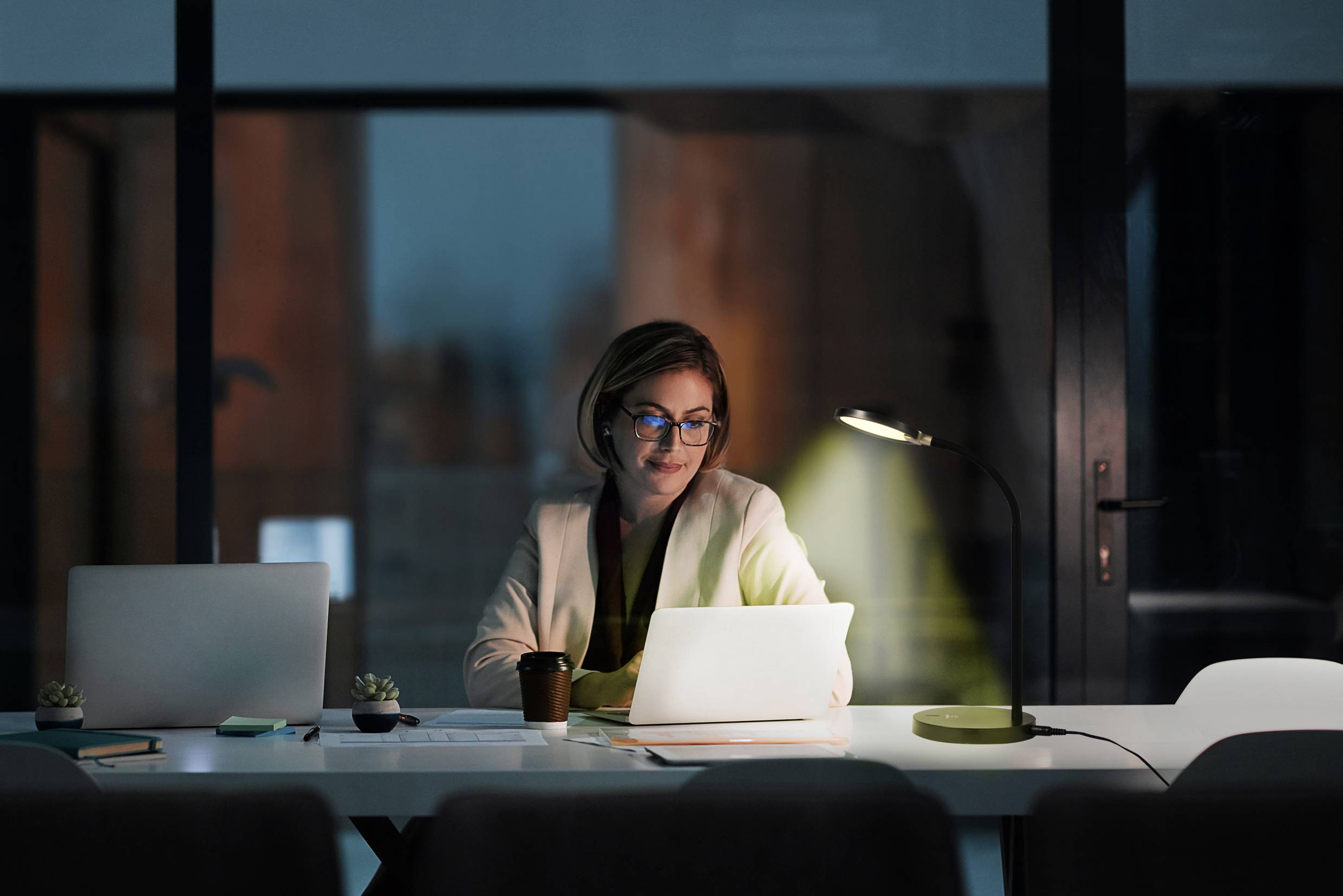 Une femme travaille le soir sur un ordinateur portable dans un bureau. La pièce est éclairée par une lampe de bureau.