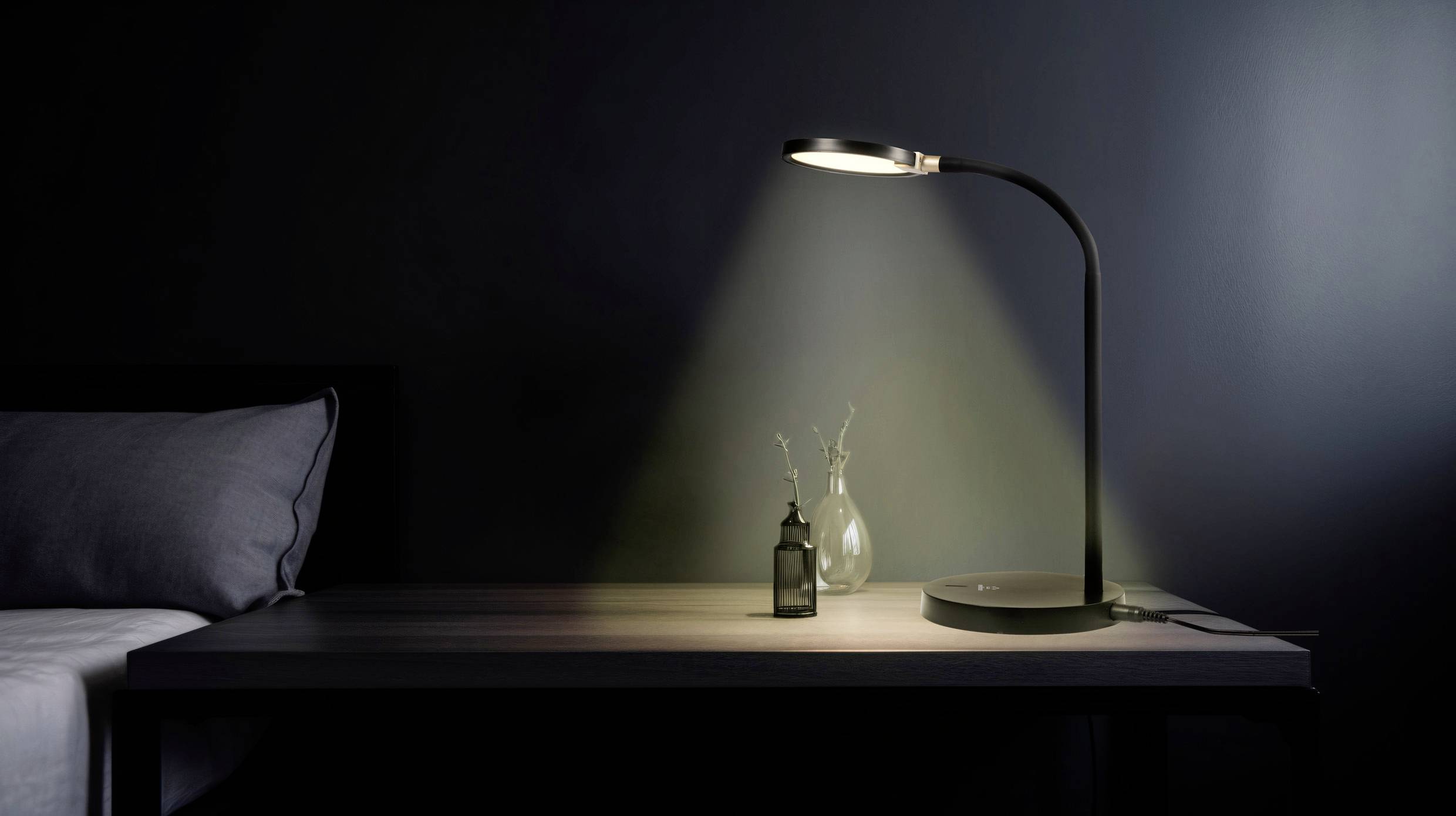 Table de nuit avec une lampe allumée projetant une lumière douce sur une table avec un petit bouquet de fleurs et une bouteille.