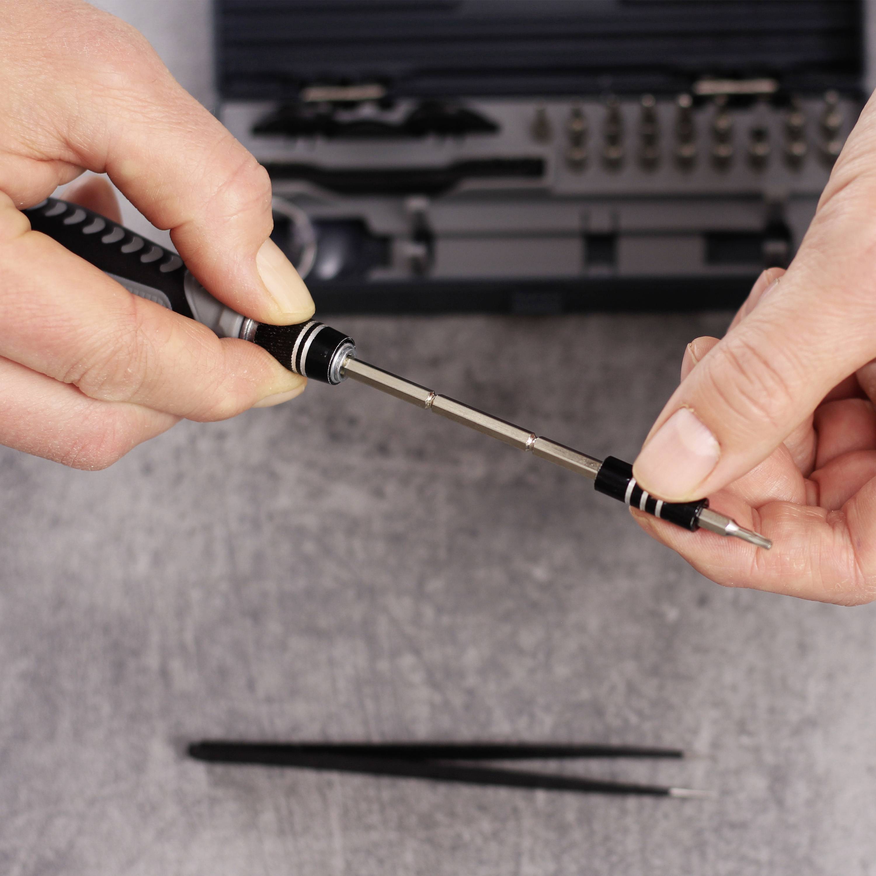 Deux mains utilisent un petit jeu de tournevis sur une surface grise, avec une boîte à outils en arrière-plan.
