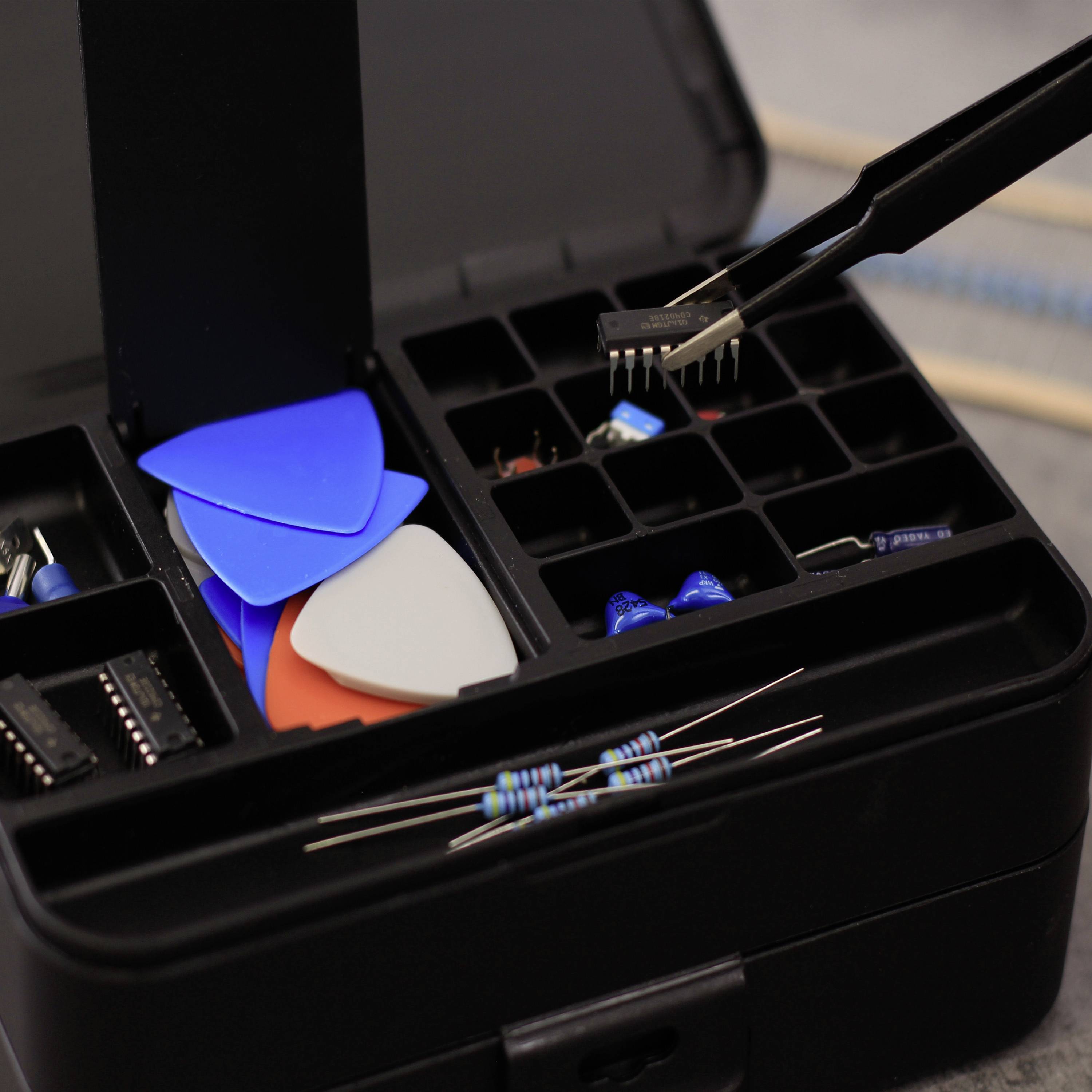 Une boîte à outils ouverte avec des accessoires électroniques : des médiators colorés, des résistances et une pince brucelles tenant une petite pièce.