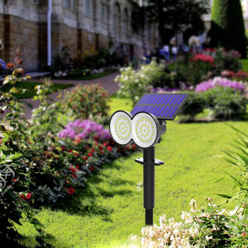 Une lampe solaire avec deux lumières LED rondes est placée dans un jardin ensoleillé devant des fleurs en fleur et des buissons verdoyants.