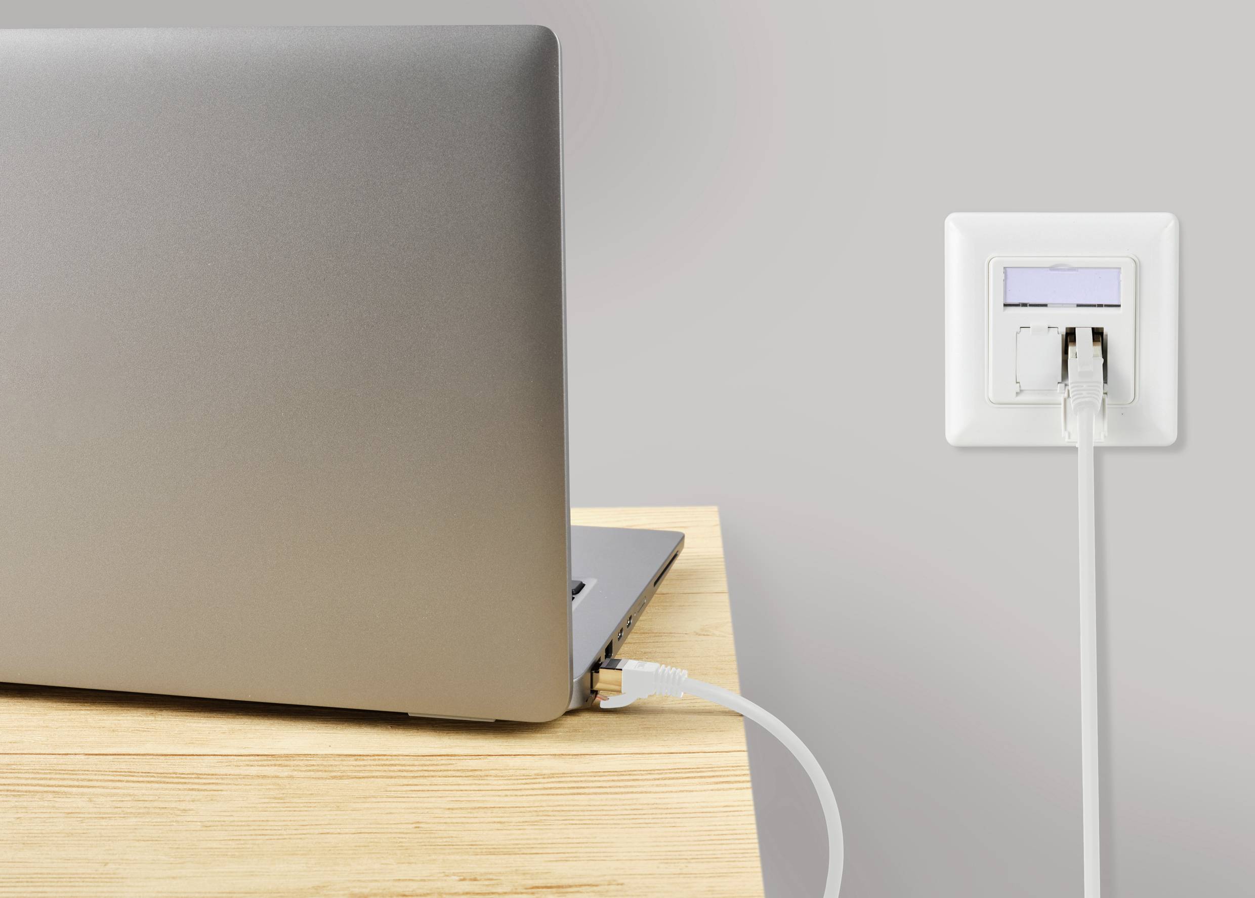 Un ordinateur portable est posé sur une table en bois et connecté à une prise murale via un câble Ethernet, transmettant un signal internet.