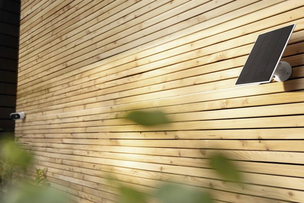 Module solaire installé sur une façade en bois, illuminé par la lumière du soleil. Une caméra est également fixée au mur.