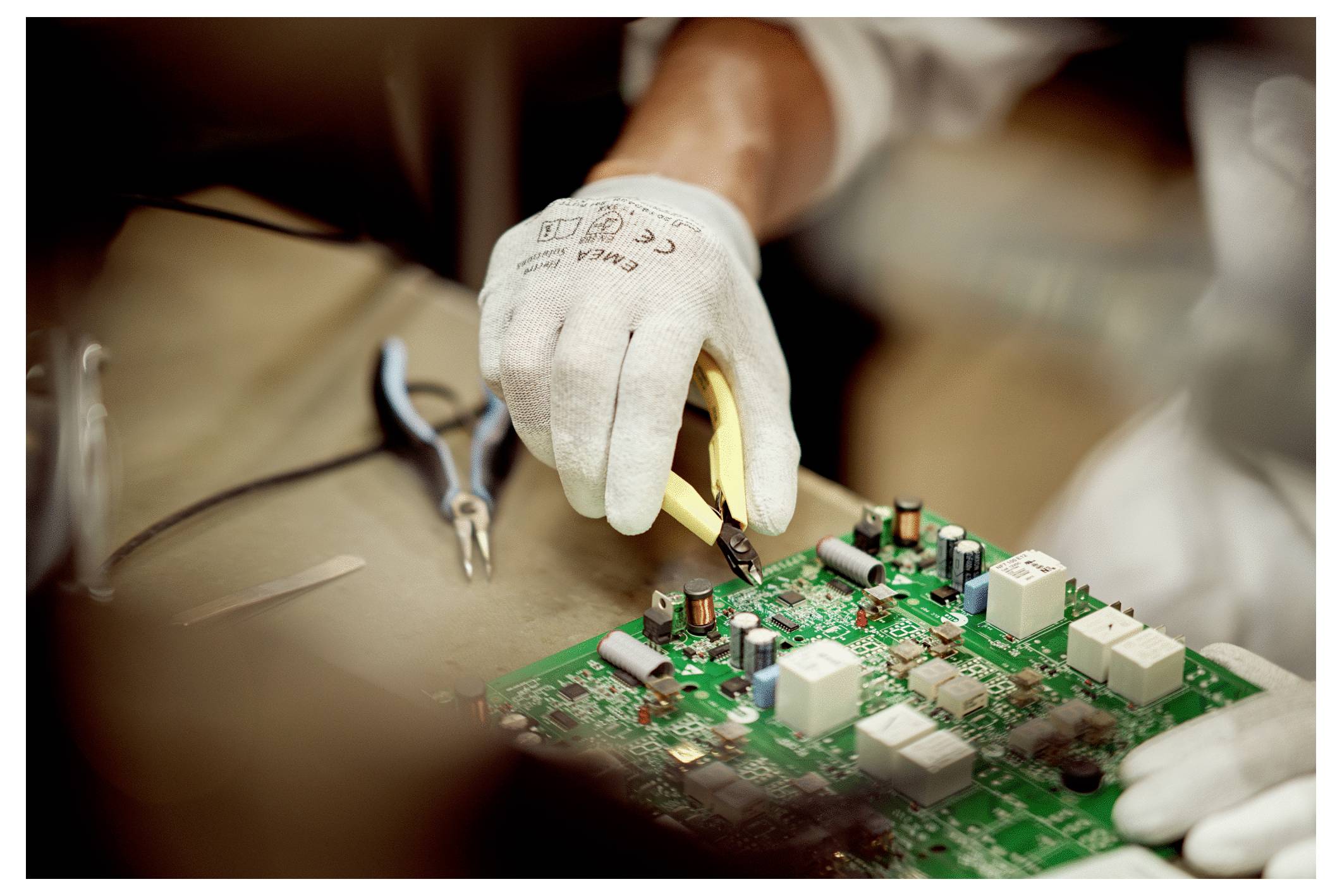 Une personne portant des gants utilise un fer à souder sur une carte de circuit imprimé verte, avec des outils comme des pinces à proximité, suggérant une concentration sur un travail électronique.